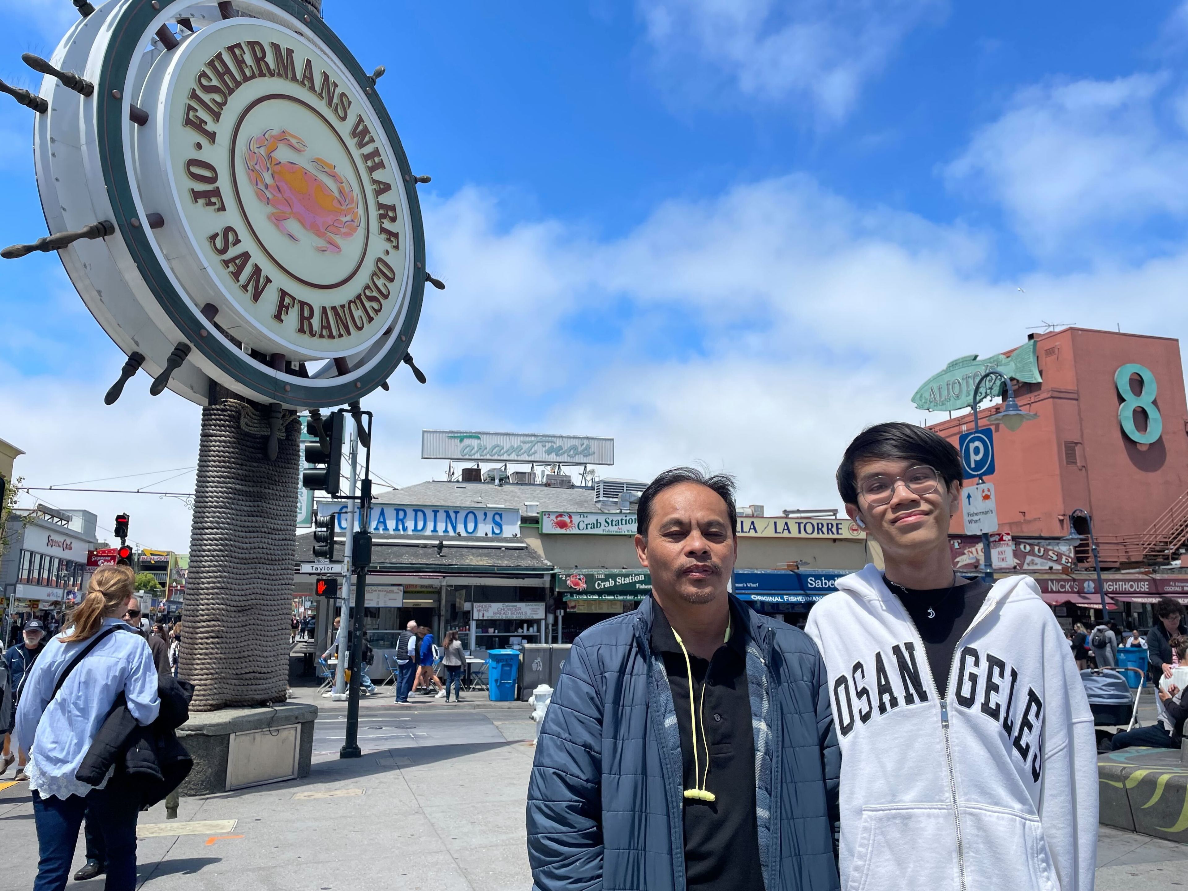 A teenage boy and a middle-aged man stand next to each other in a bustling shopping area for a photo.