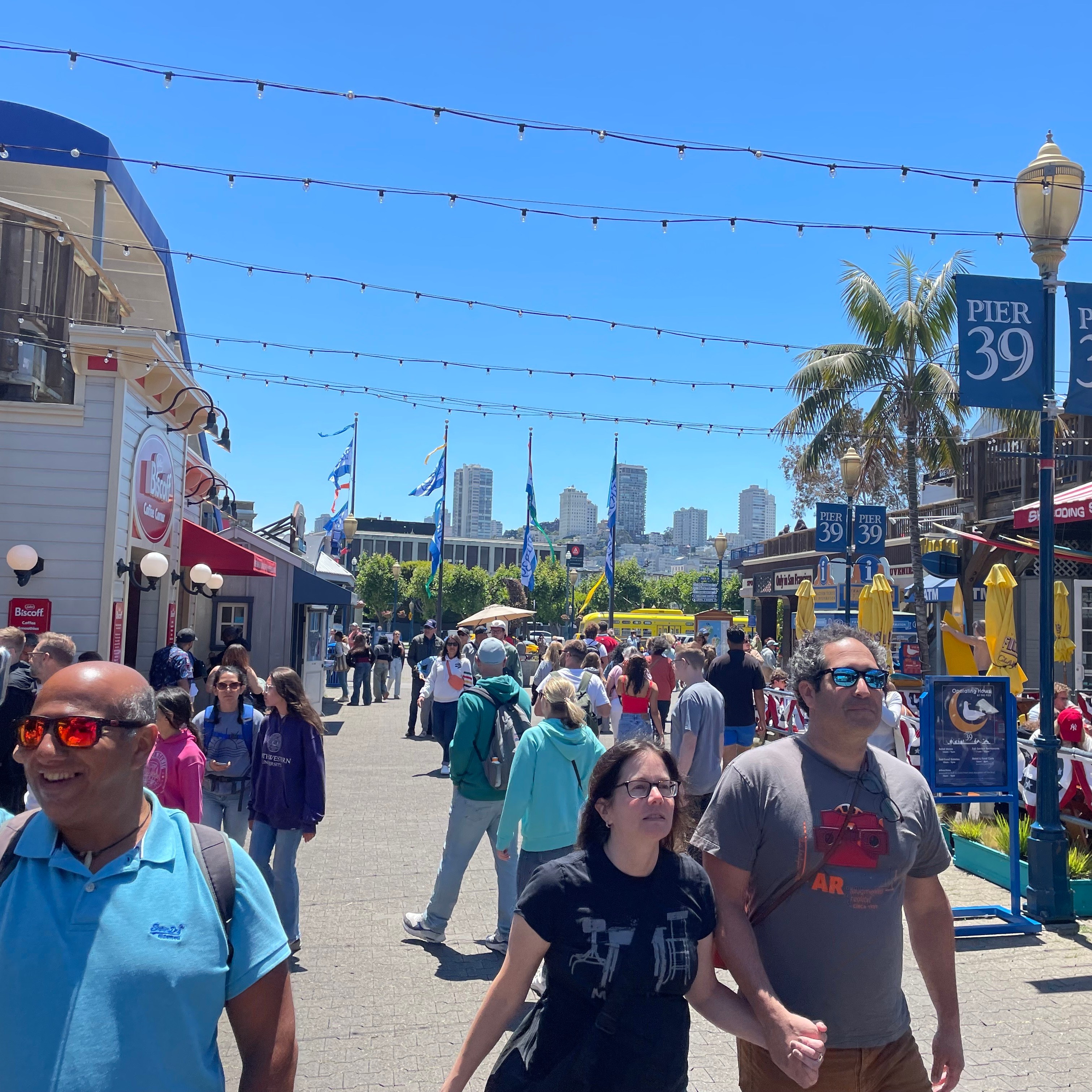 People stroll along a busy, sunlit Pier 39, with colorful storefronts, flags, and a slight urban skyline in the background; one person is taking a selfie.