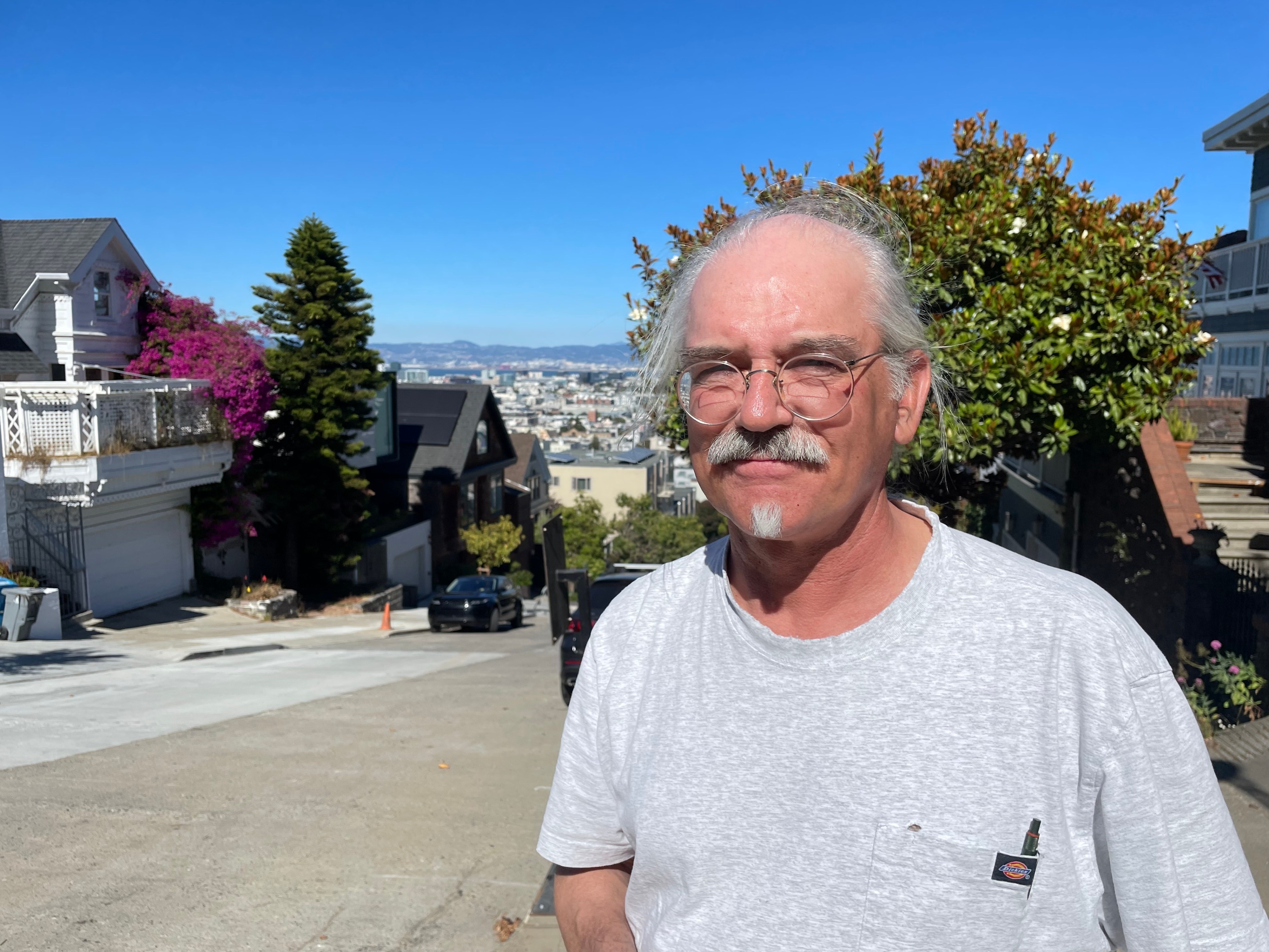 A man with glasses and a mustache stands on a sunny residential street with houses, greenery, and a clear blue sky in the background.