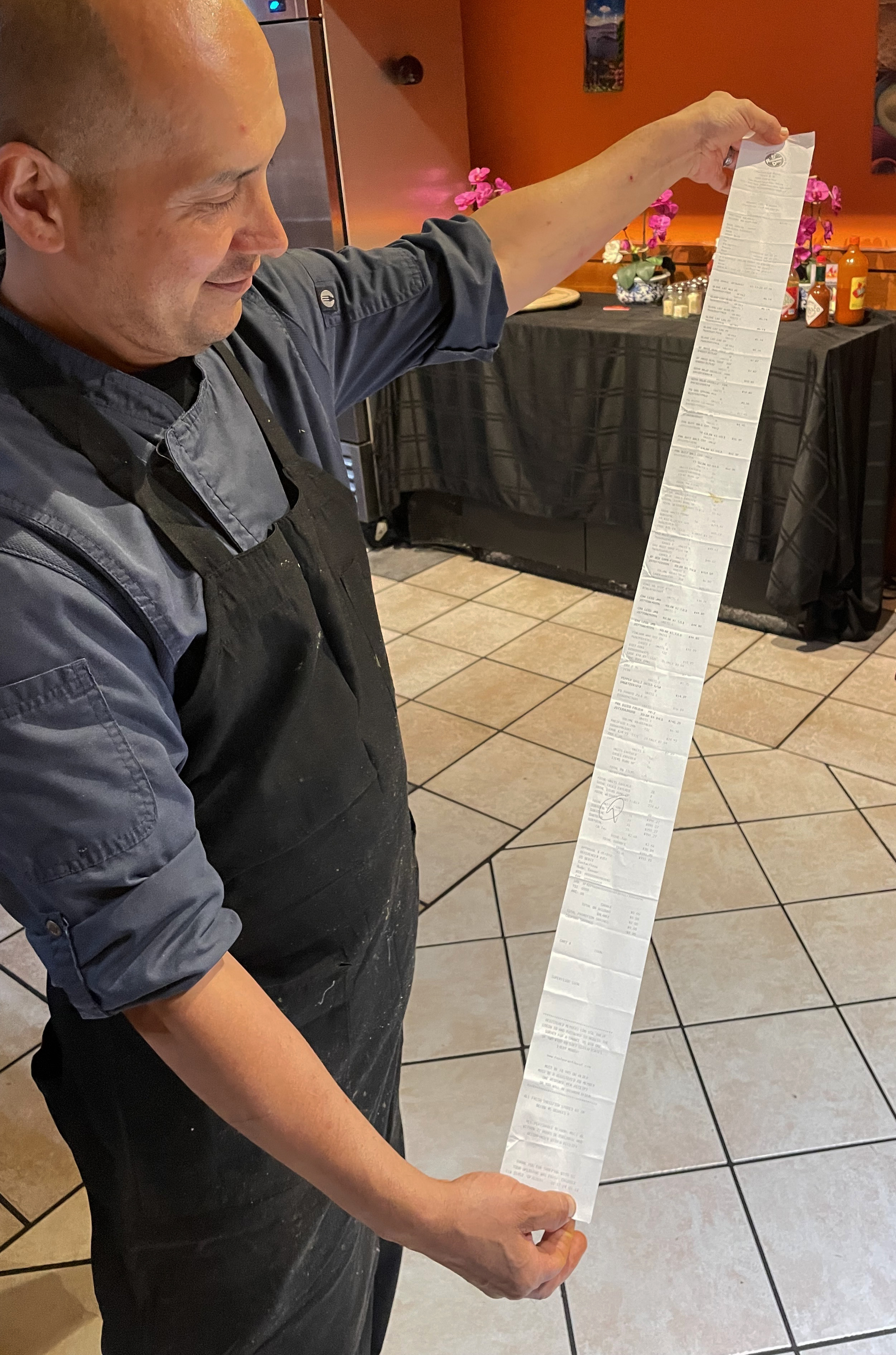 A person in a dark apron and blue shirt is holding a very long receipt, extending it from above their head to their waist. The background has orange walls and a table with items.