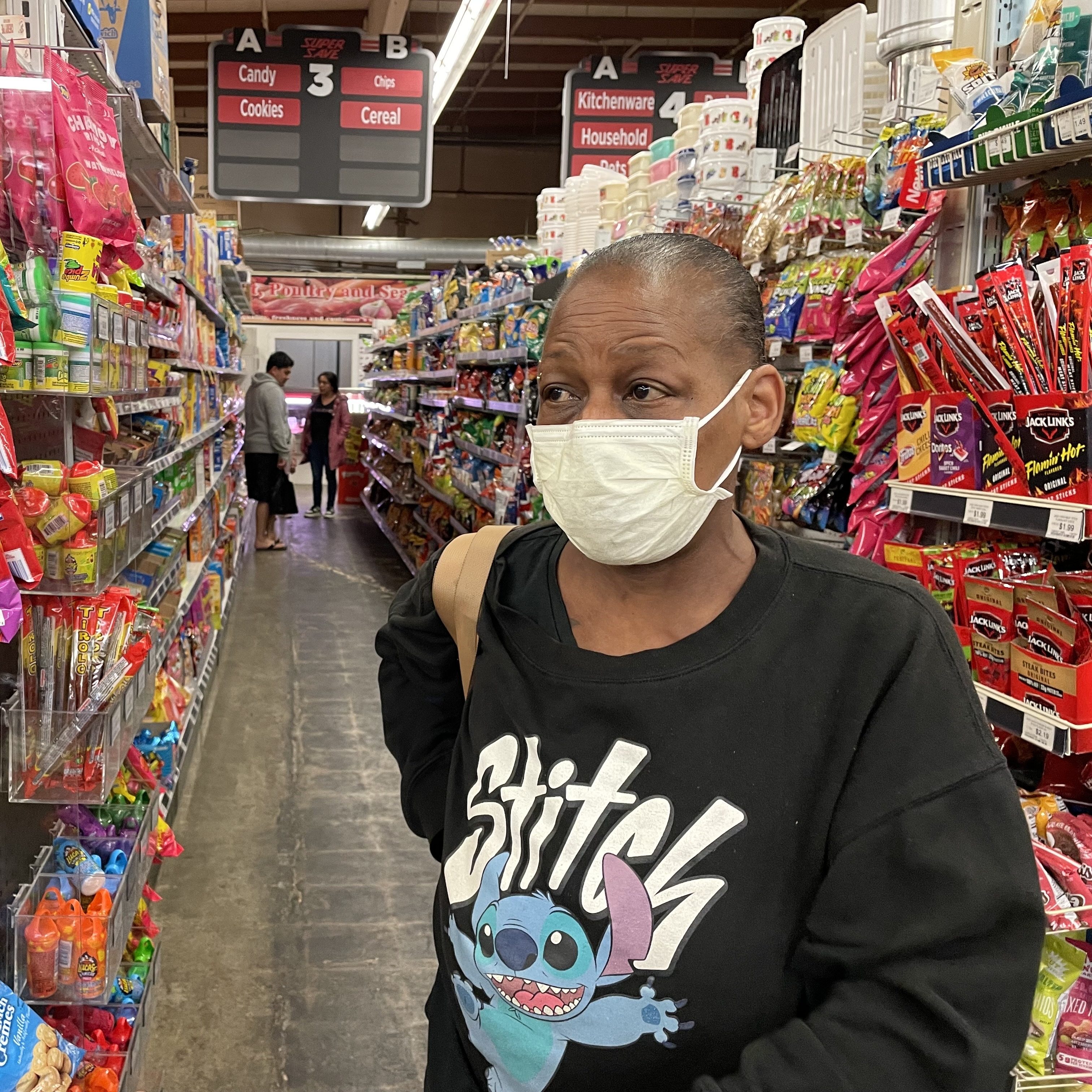 A masked woman in a Stitch shirt stands in a grocery aisle filled with colorful snacks and candies; other shoppers and signs are visible in the background.