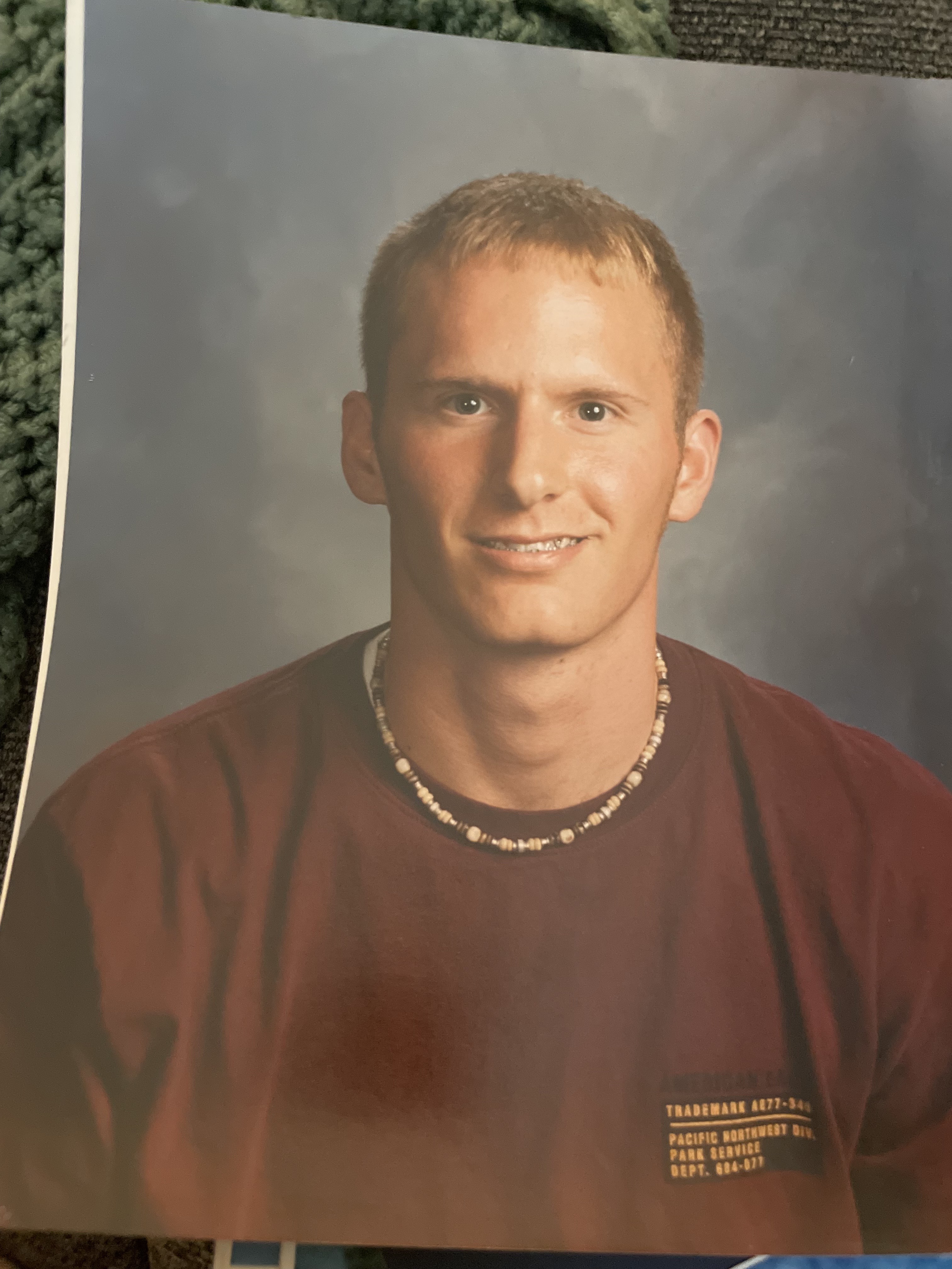 A young man with short blond hair wears a beaded necklace and a maroon T-shirt. He smiles slightly against a simple, muted backdrop.