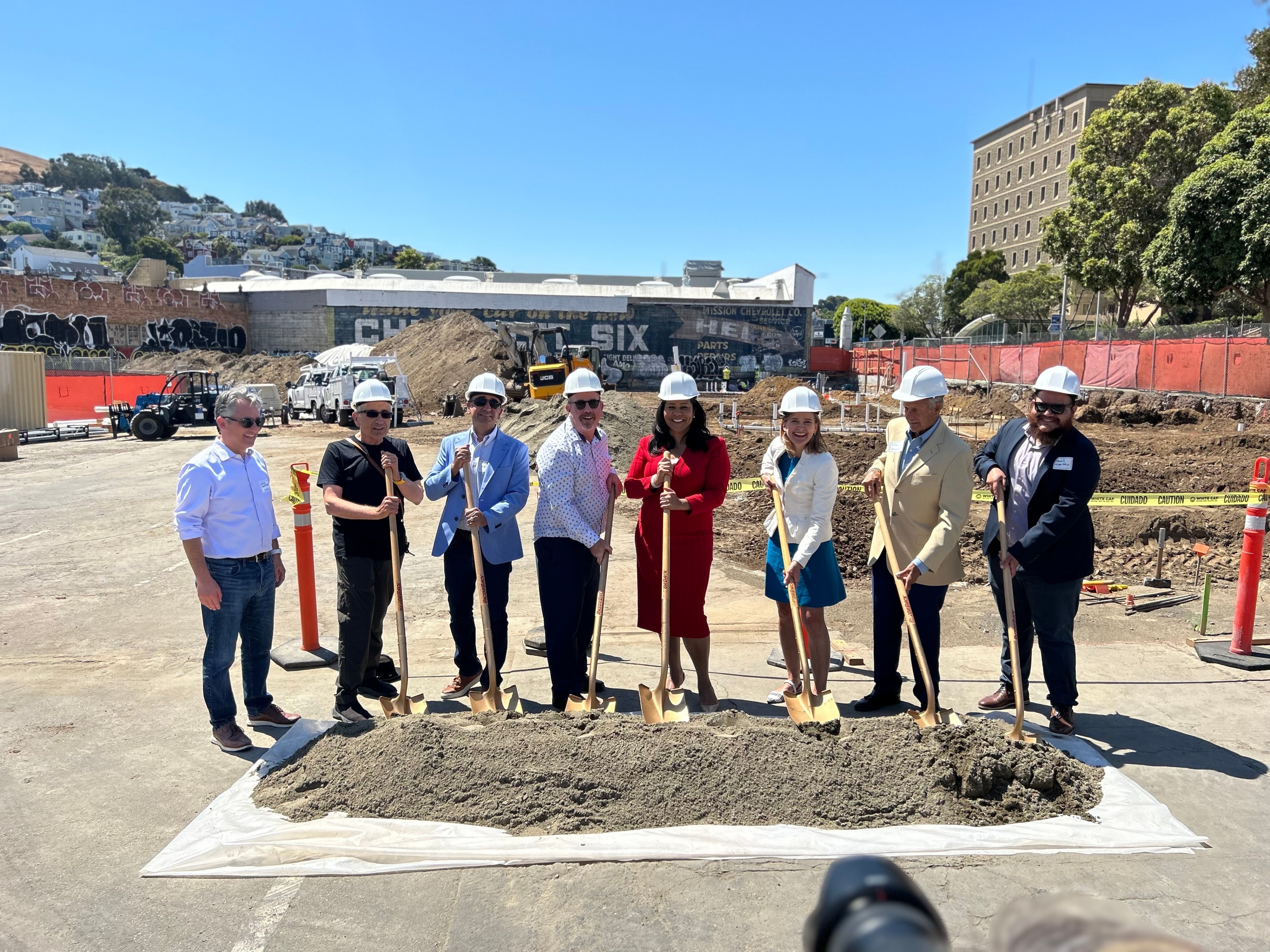 A group of eight people in hard hats stand at a construction site, posing with shovels over a mound of dirt, with buildings and heavy machinery in the background.