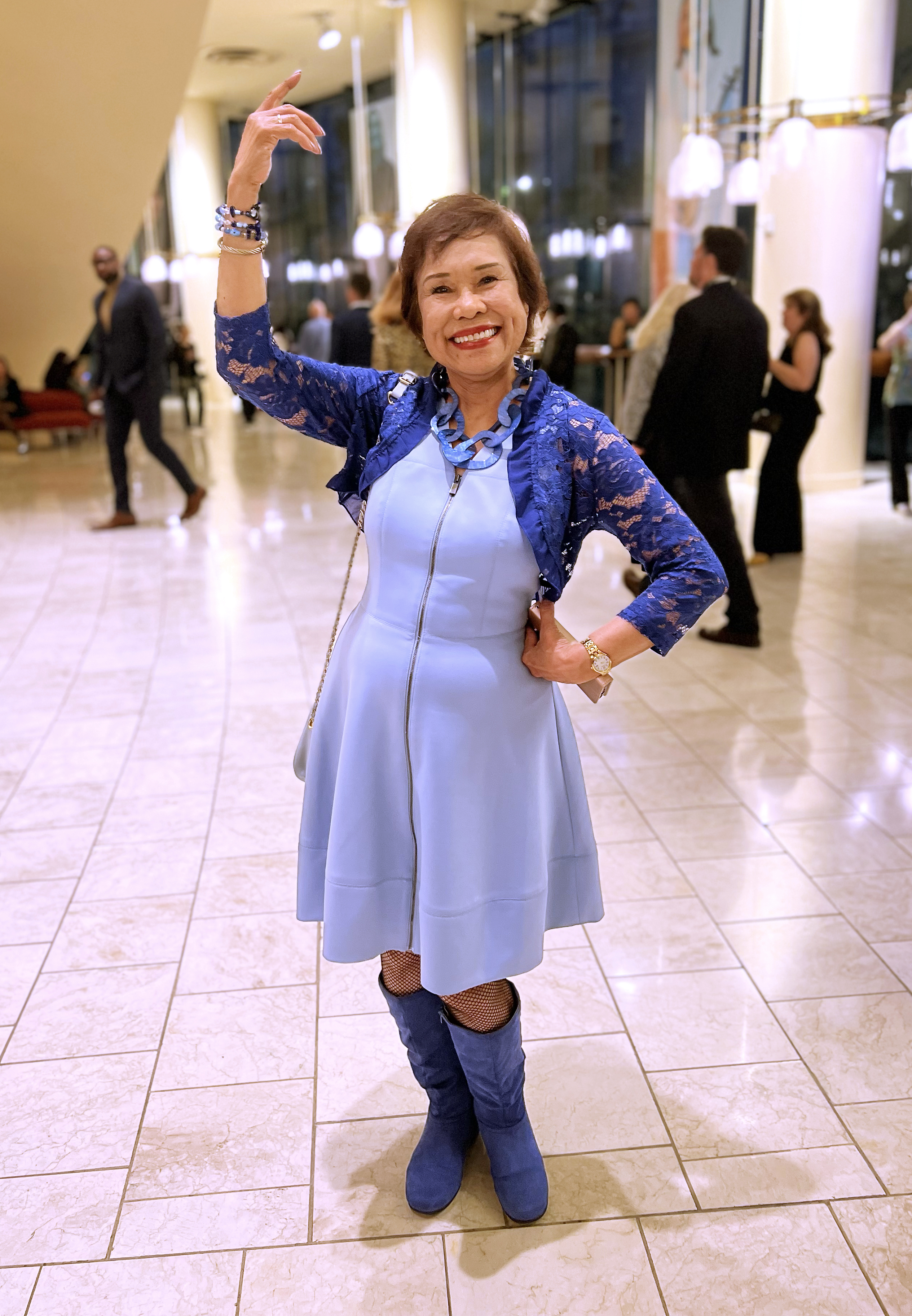 A joyful woman in a light blue dress and matching blue boots poses inside a well-lit, elegant venue with marble floors, raising one arm gracefully while smiling broadly.