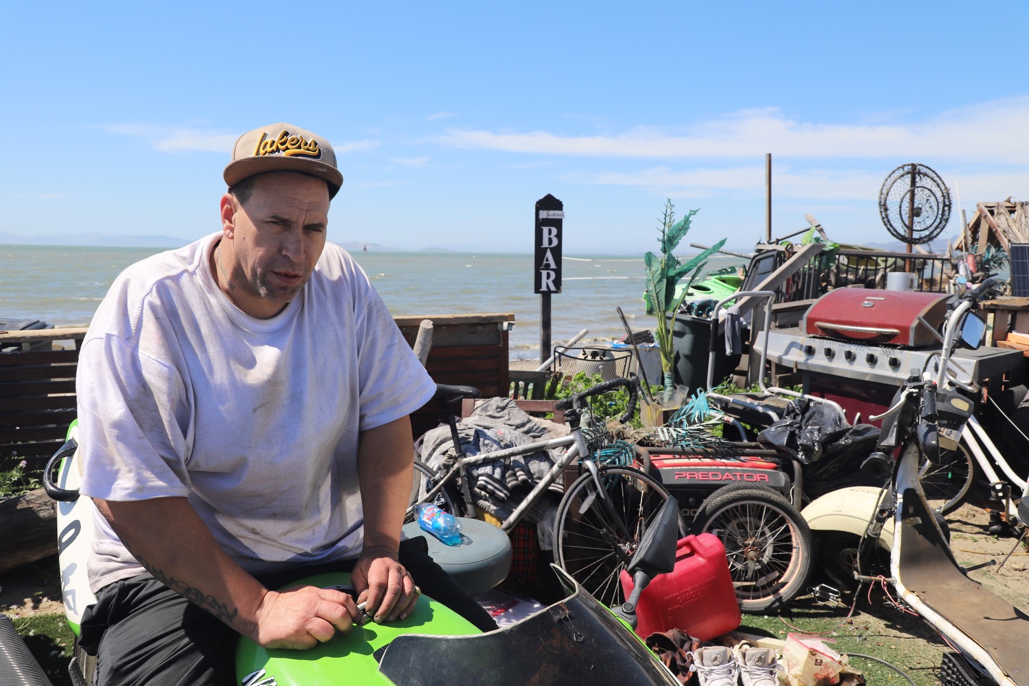 A man in a white shirt and cap is sitting outside near miscellaneous items like a barbecue grill and bikes, with the ocean and a &quot;BAR&quot; sign in the background.