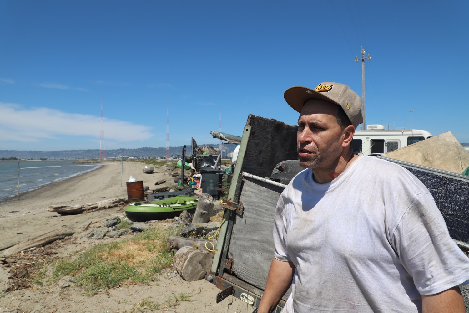 A man in a white shirt and beige cap stands on a cluttered sandy shore beside a makeshift shelter. The background shows a bay, towers, and distant hills.