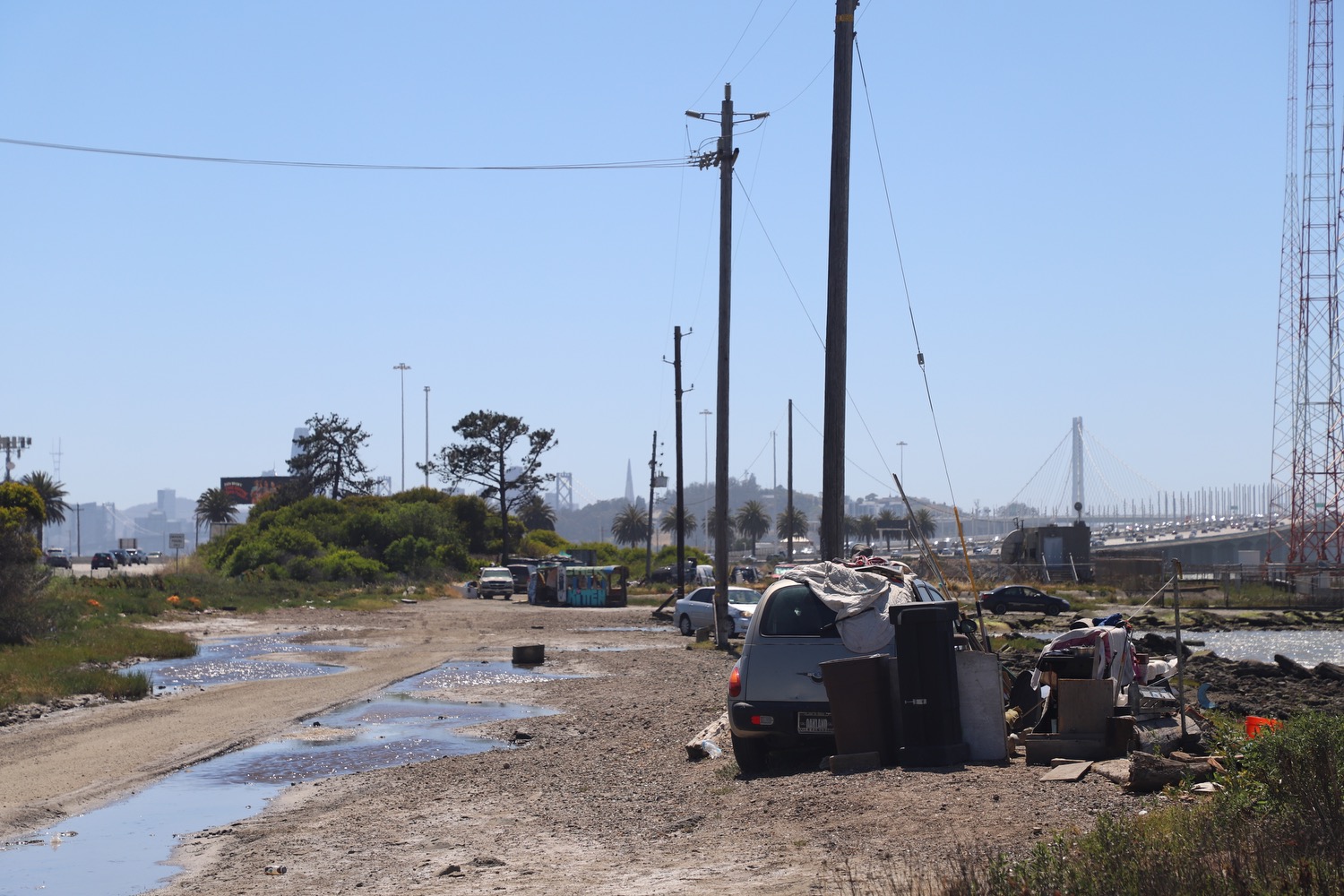 The image shows a muddy dirt road lined with utility poles. There are scattered vehicles and various items, leading to a bridge seen in the distance under a clear sky.