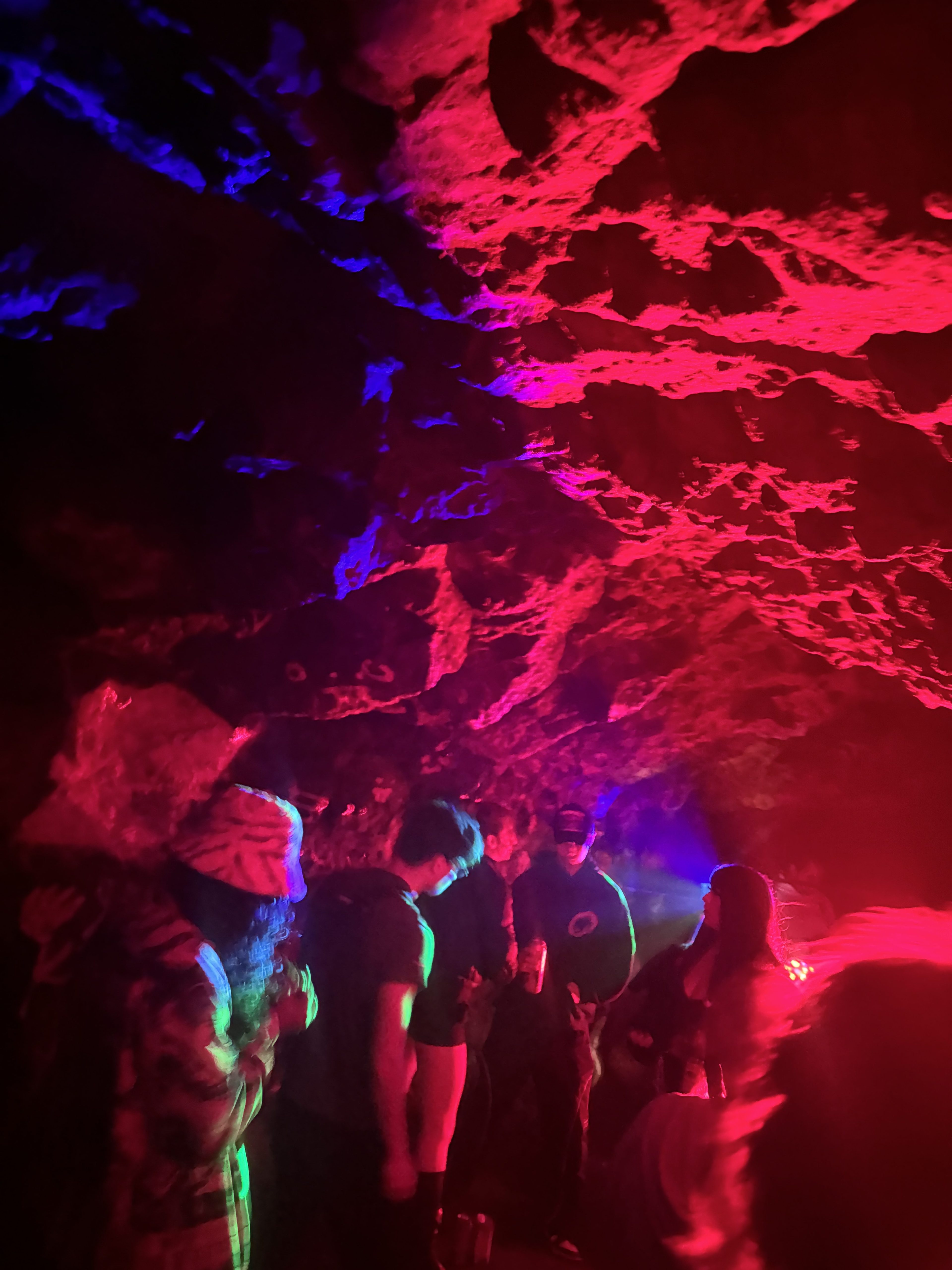 People are gathered in a dimly lit cave decorated with colorful red and blue lights casting shadows on the rocky ceiling, creating a vibrant, lively atmosphere.