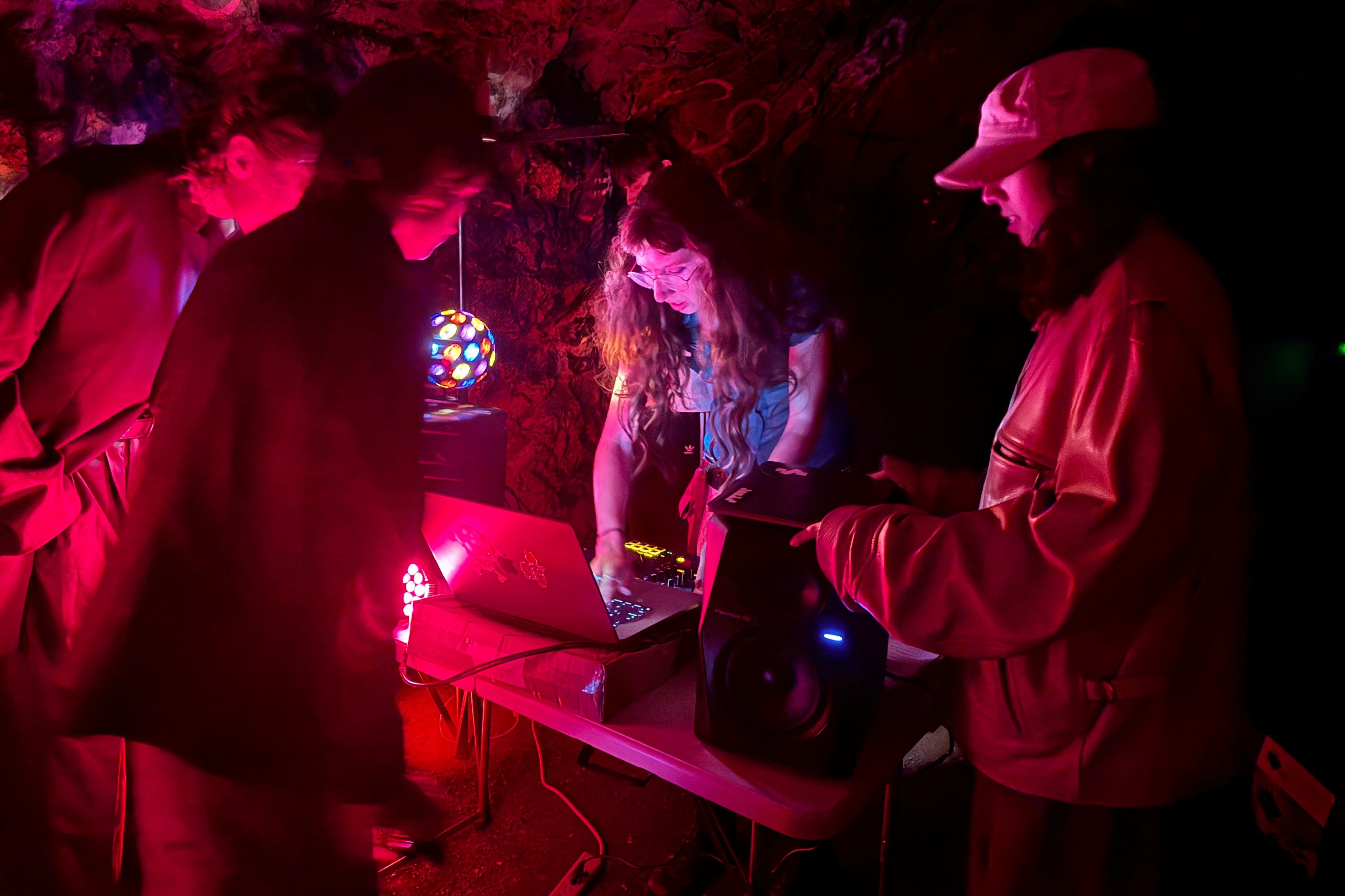 A dimly lit cave features four people gathered around electronic equipment on a table, illuminated by colorful lights, including a disco ball and a glowing red light.