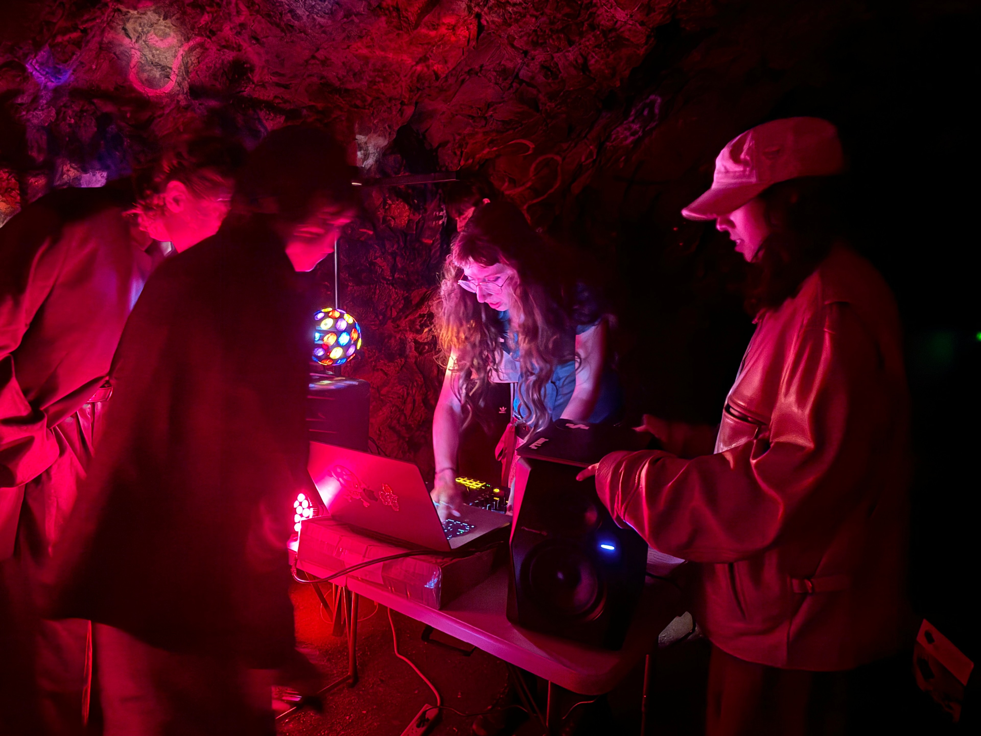 A dimly lit cave features four people gathered around electronic equipment on a table, illuminated by colorful lights, including a disco ball and a glowing red light.