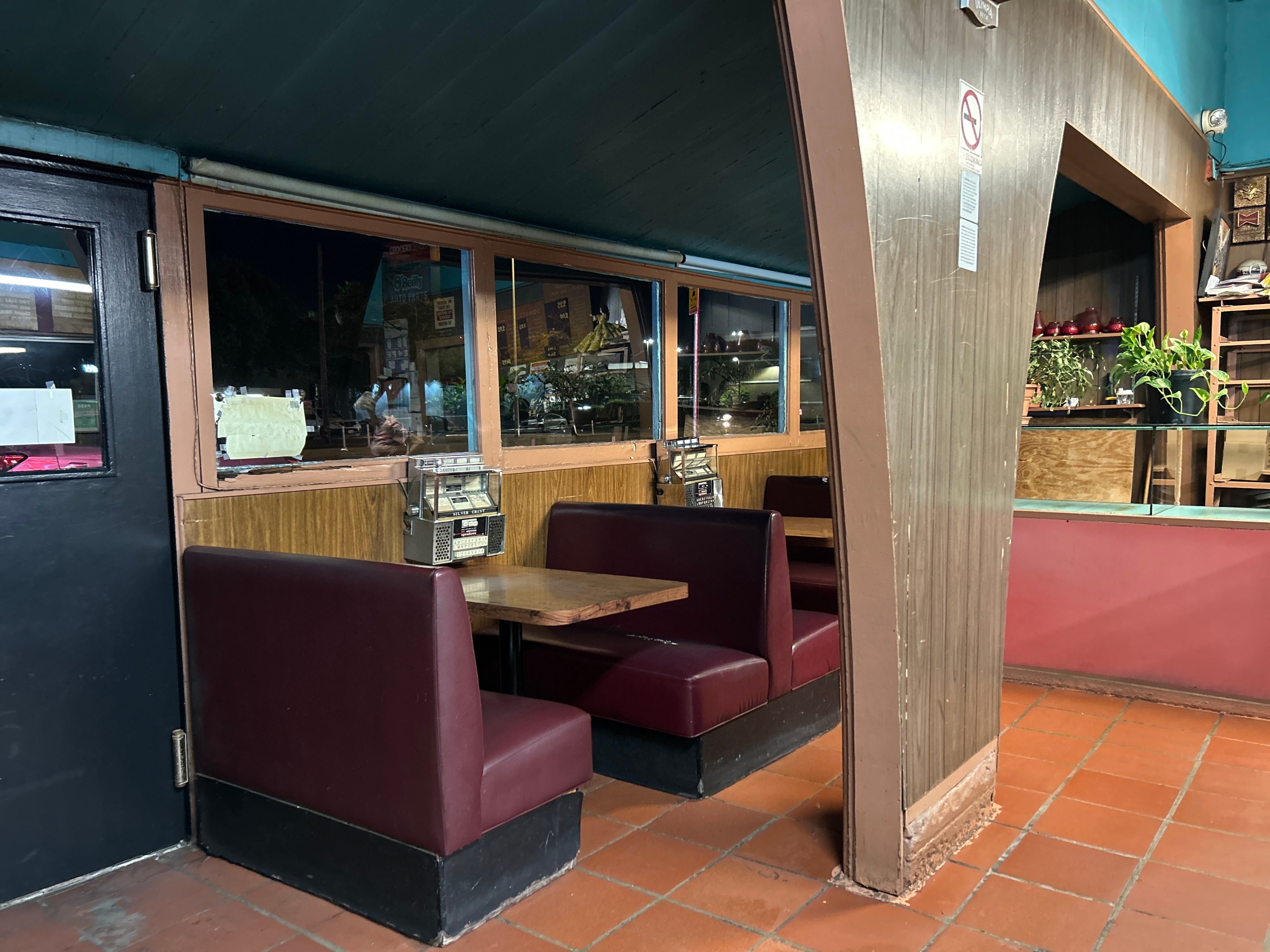 The image shows a retro diner with a booth that has red seats, a wooden table, and a small jukebox. There are potted plants and ornaments on nearby shelves.