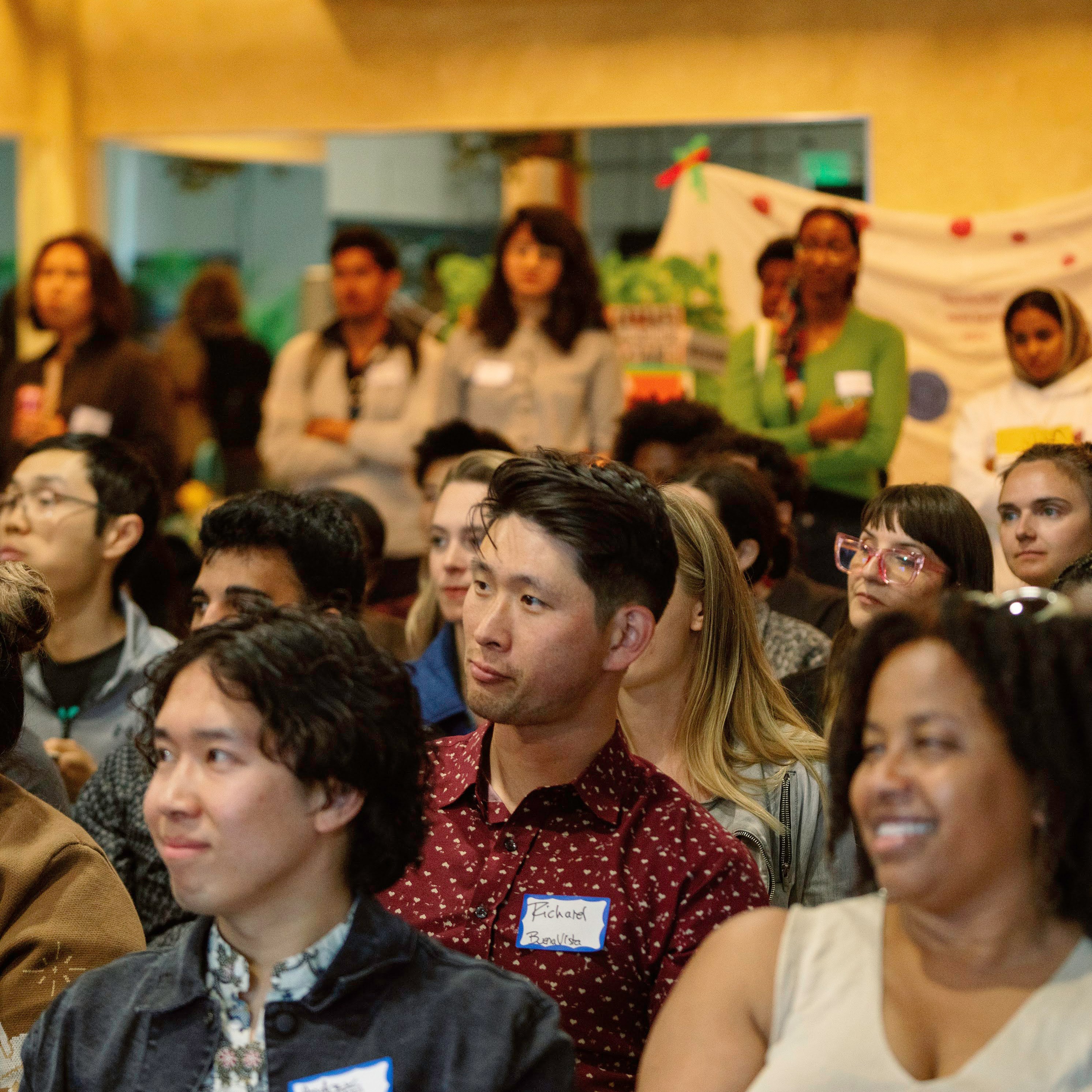 A diverse group of people attentively watch something out of the frame in an indoor setting, with a mix of sitting and standing individuals. Some wear name tags.