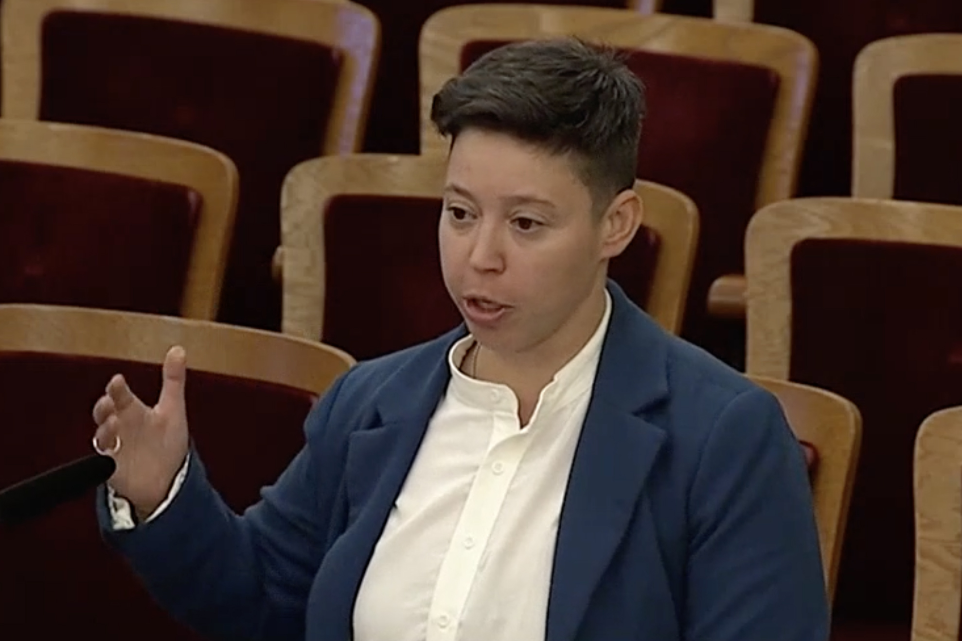 A person with short hair is speaking animatedly in a lecture hall, wearing a white shirt and blue blazer against a backdrop of empty, red-cushioned wooden chairs.