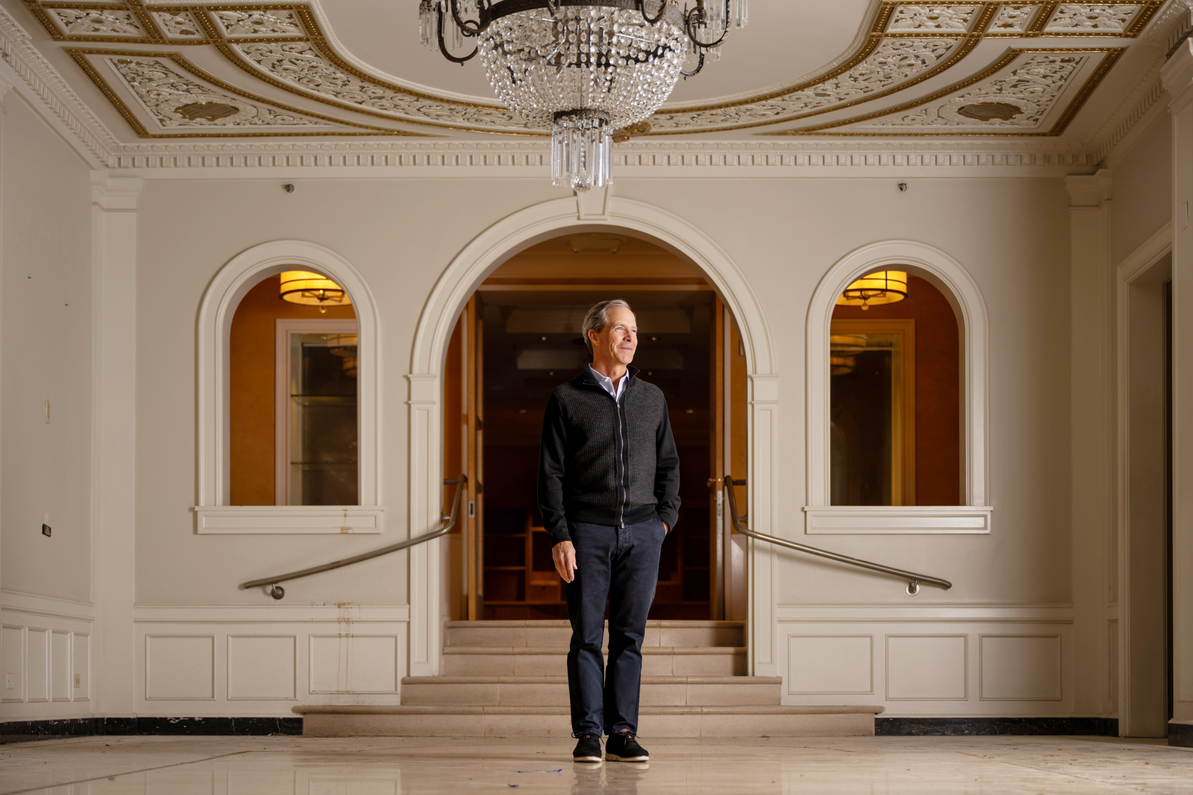 A person in a dark sweater stands in a brightly lit, ornate room with white walls, arched doorways, a chandelier, and decorative ceiling details.