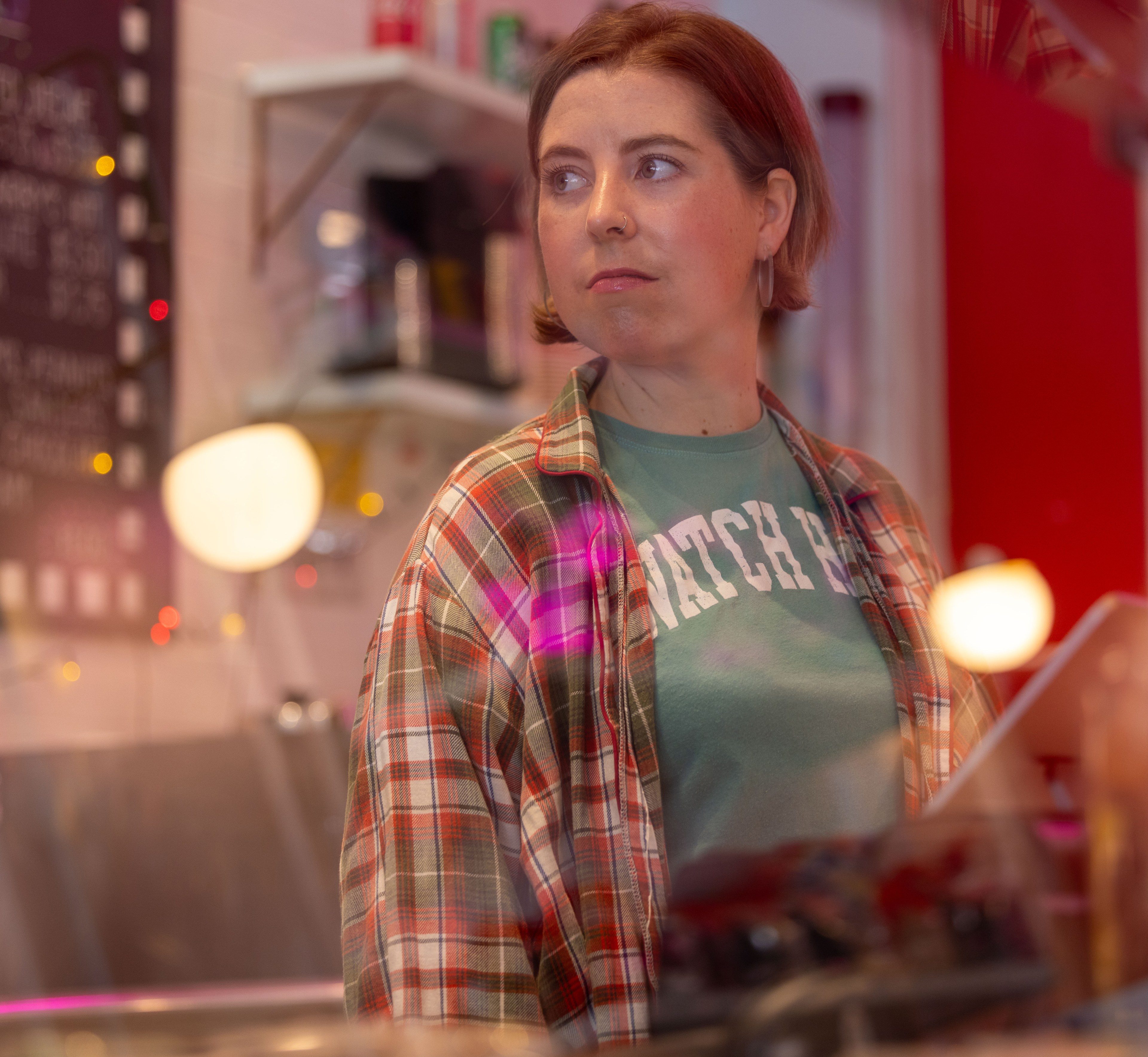 A woman with short hair looks off-camera in a cozy, dimly lit setting. She wears a green shirt and a plaid jacket. String lights and shelves are visible in the background.