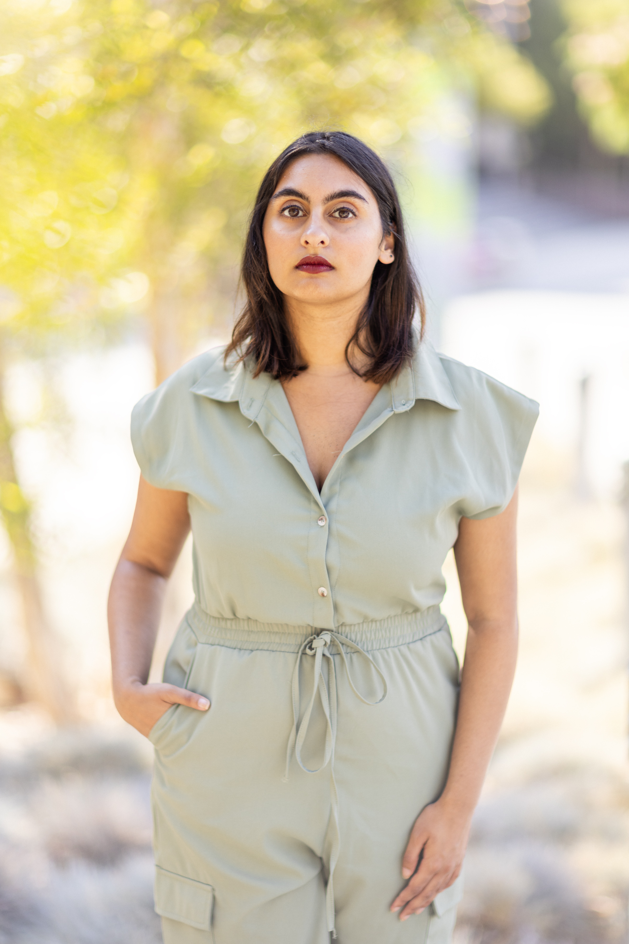 Zahra Hajee stands outdoors in a light sage green jumpsuit with her right hand in a pocket and an arm relaxed at her side. She looks directly at the camera, and the background is softly blurred.