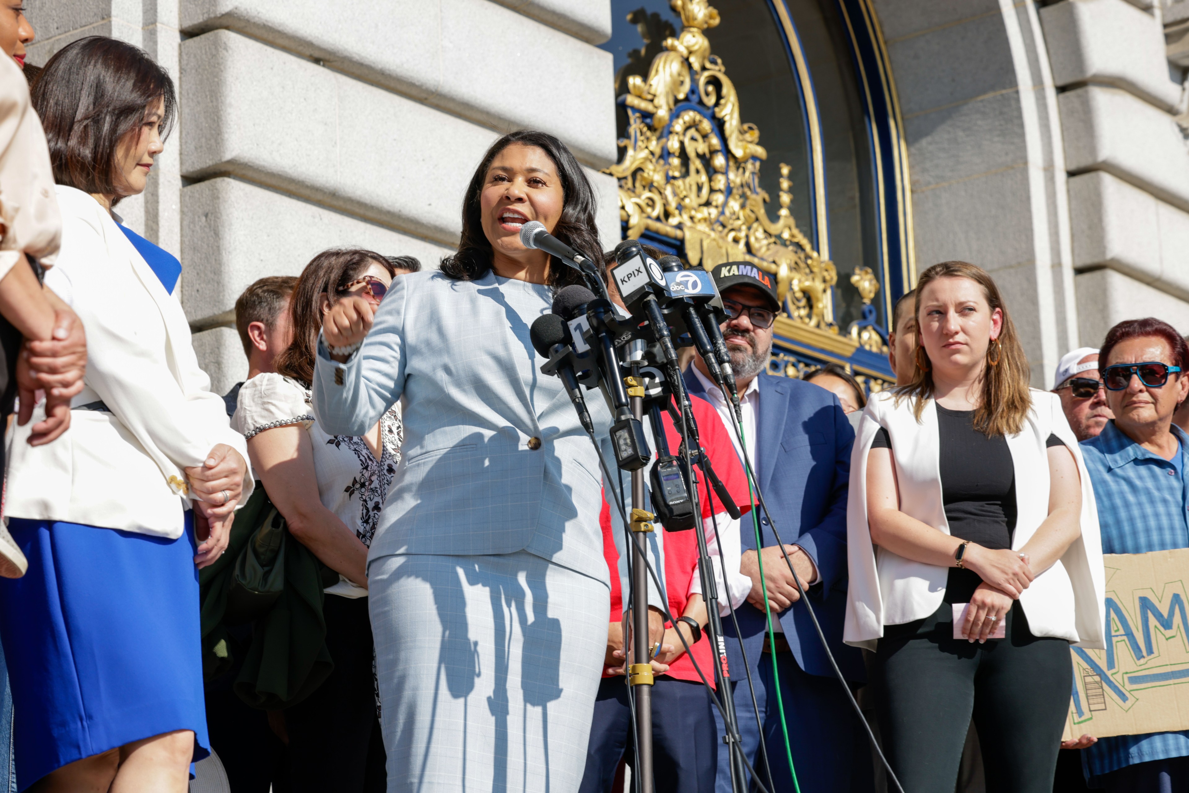 A woman in a light blue suit speaks passionately at multiple microphones, surrounded by supporters in business and casual attire, against a grand building backdrop.