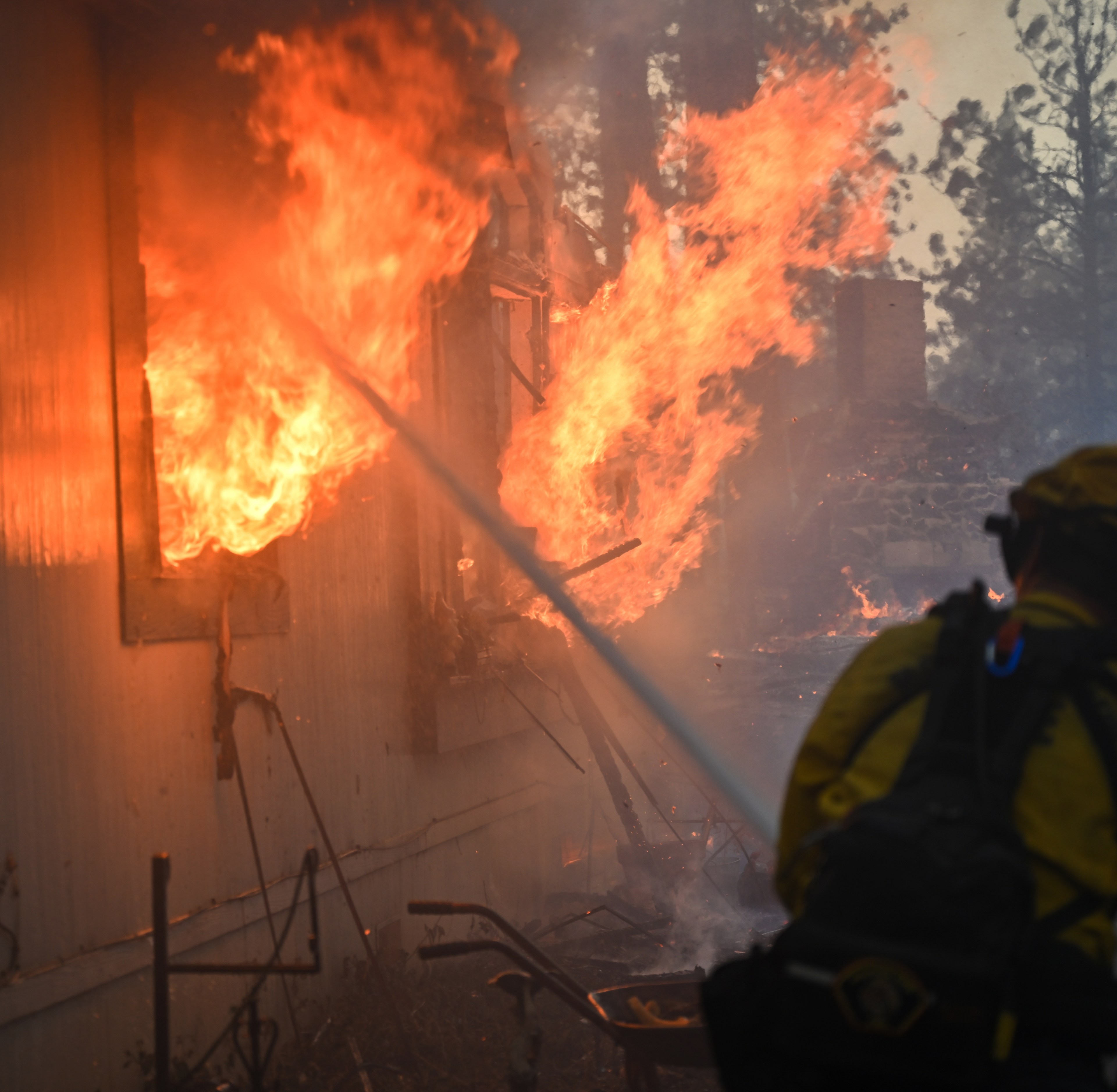 A firefighter in yellow gear is battling intense flames engulfing a building, spraying water to control the fire amidst a smoky, forested setting.