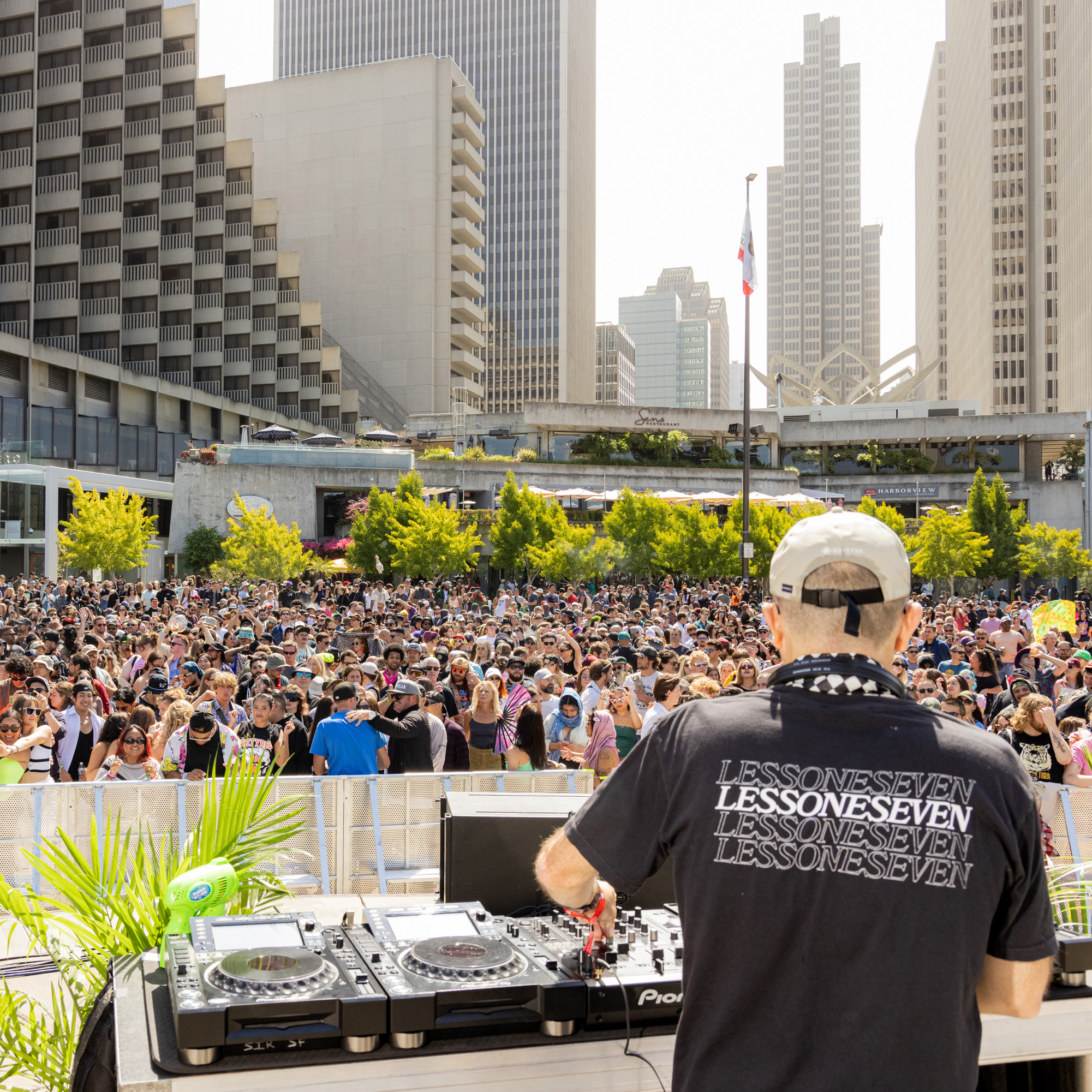 A DJ performs to a large crowd in an outdoor urban setting with tall buildings in the background. The audience is densely packed, enjoying the music in bright sunlight.