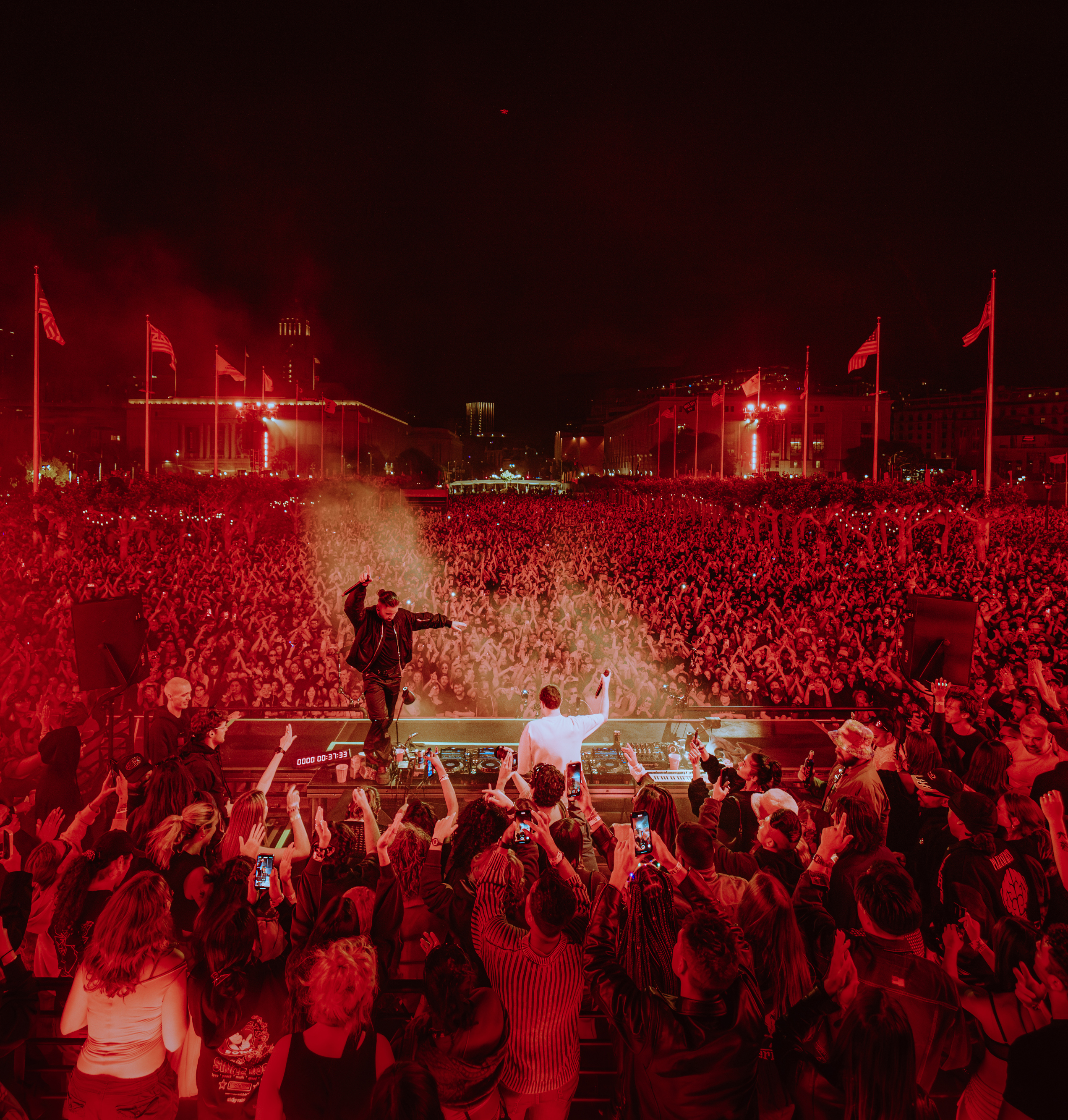 A DJ performs on stage, illuminated by red lights, in front of a massive crowd at night. The audience, packed tightly, raises their hands and phones, capturing the moment.