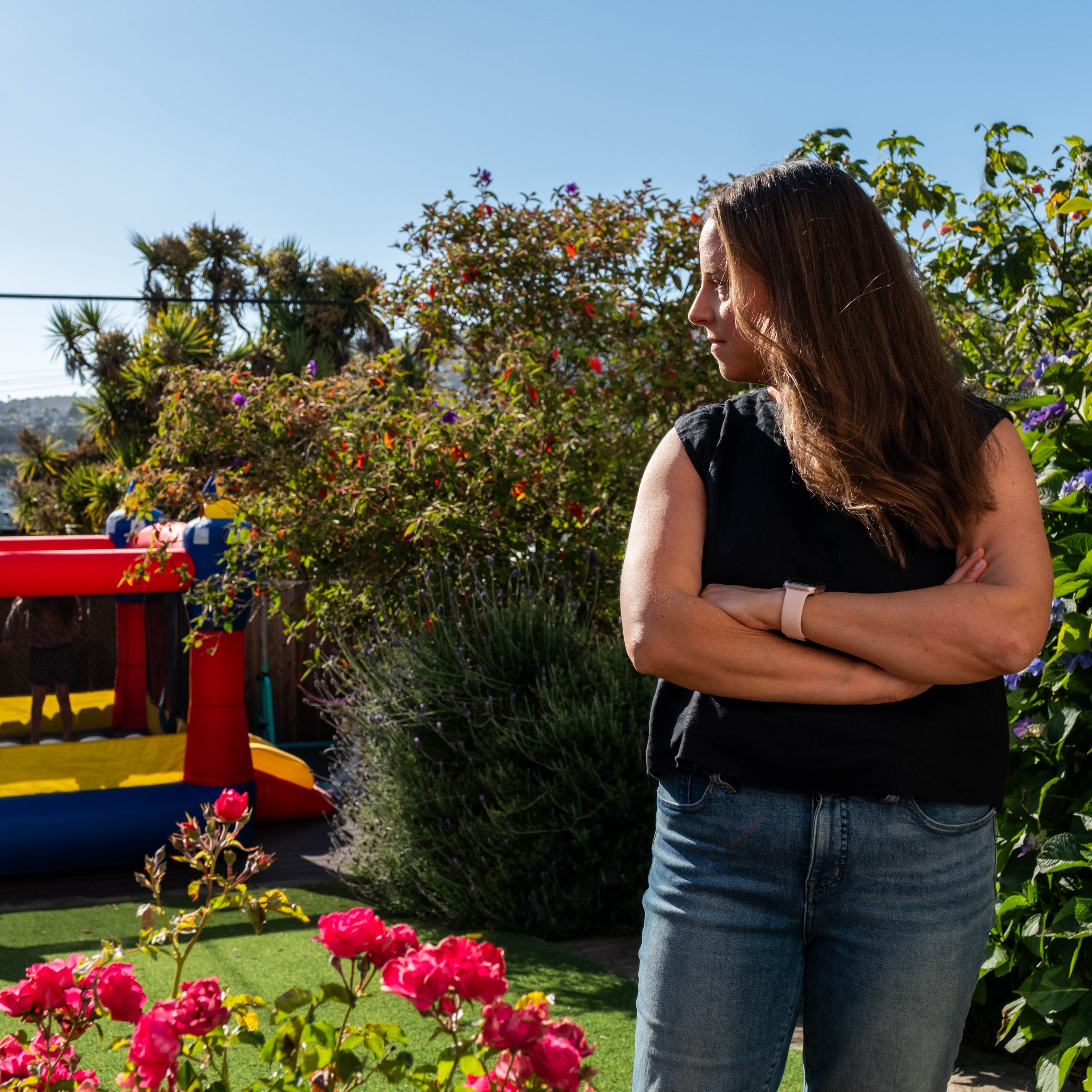 A woman stands with arms crossed in a sunny garden with pink flowers. Behind her is a red, blue, and yellow bouncy house and lush green plants.