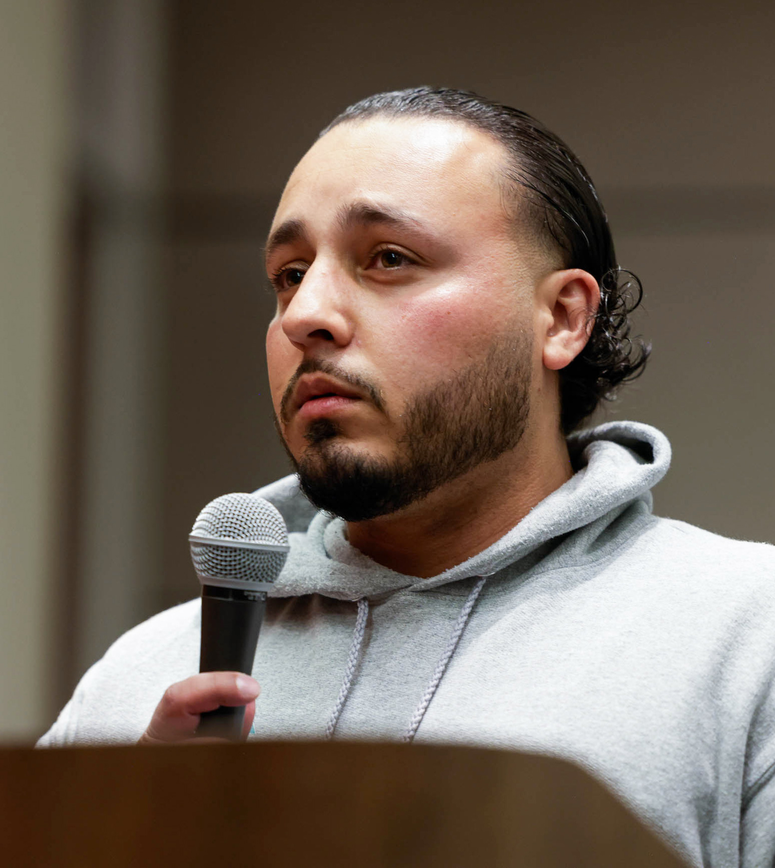 Kevin Ortiz with short, slicked-back hair and a beard is speaking into a microphone while wearing a gray hoodie. He appears to be indoors, with a neutral background.