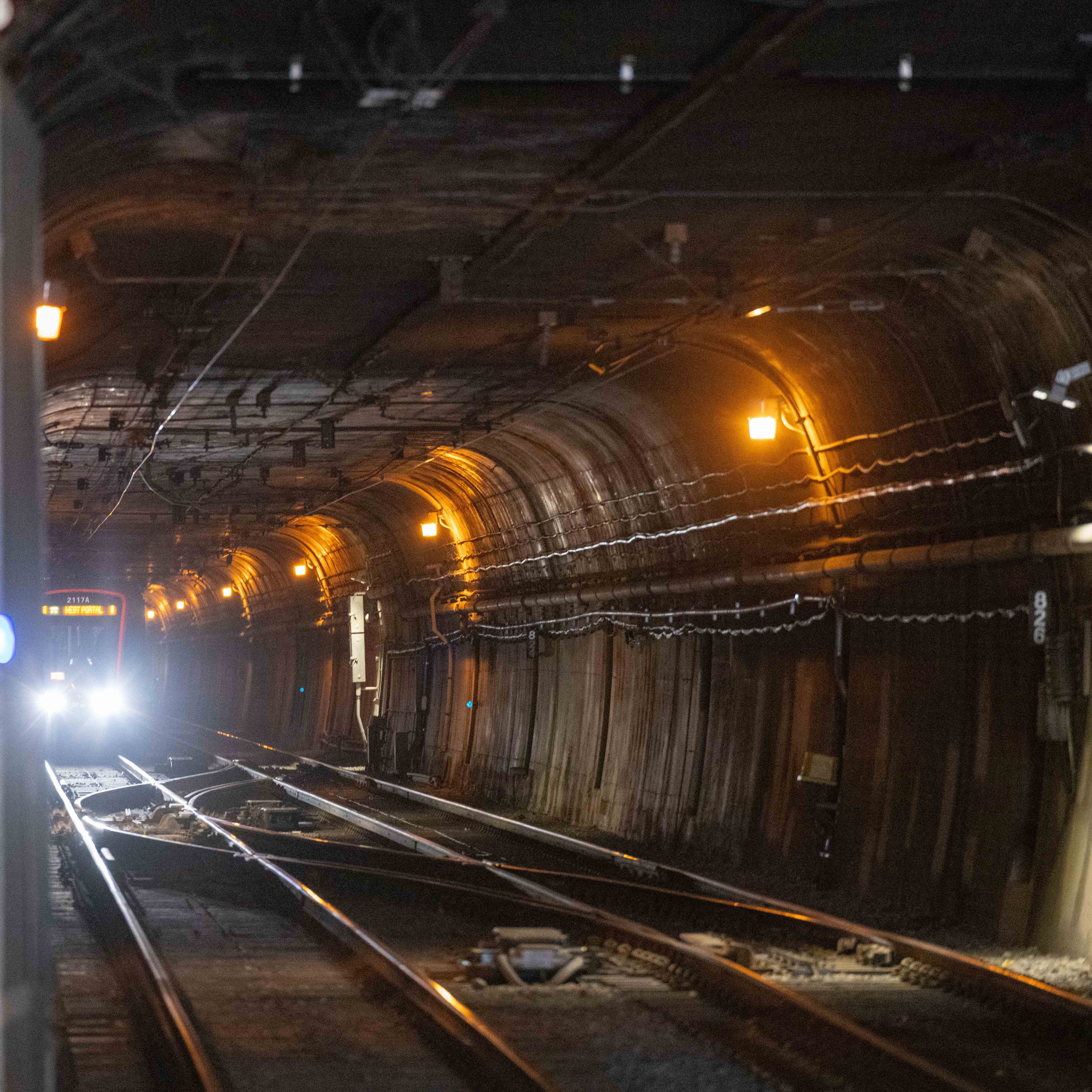 A dimly lit subway tunnel with illuminating orange lights shows train tracks curving and converging. A train with bright headlights is approaching in the distance.