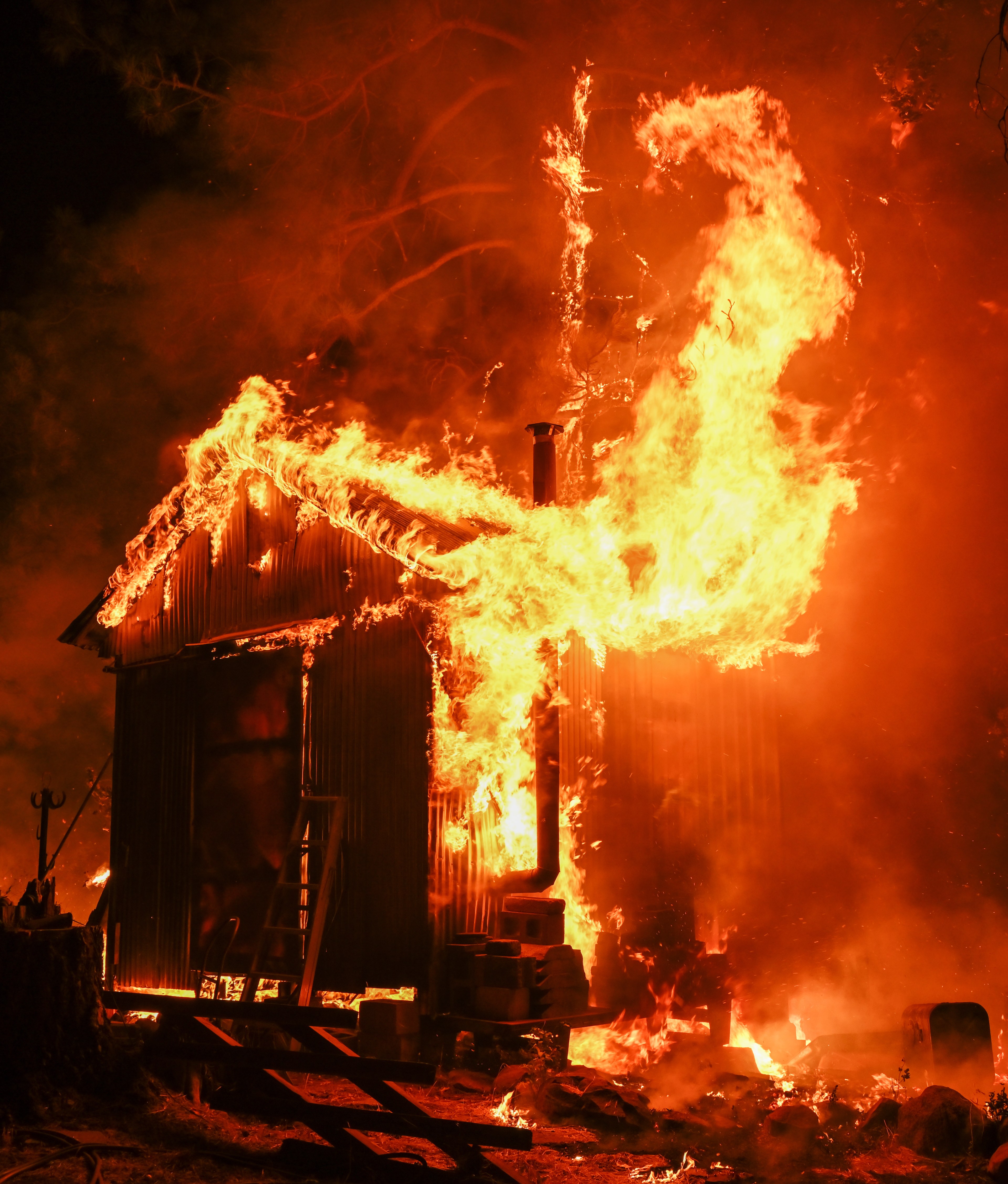 A small wooden structure is engulfed in bright orange flames, with fire consuming the roof and walls, illuminating the dark night.