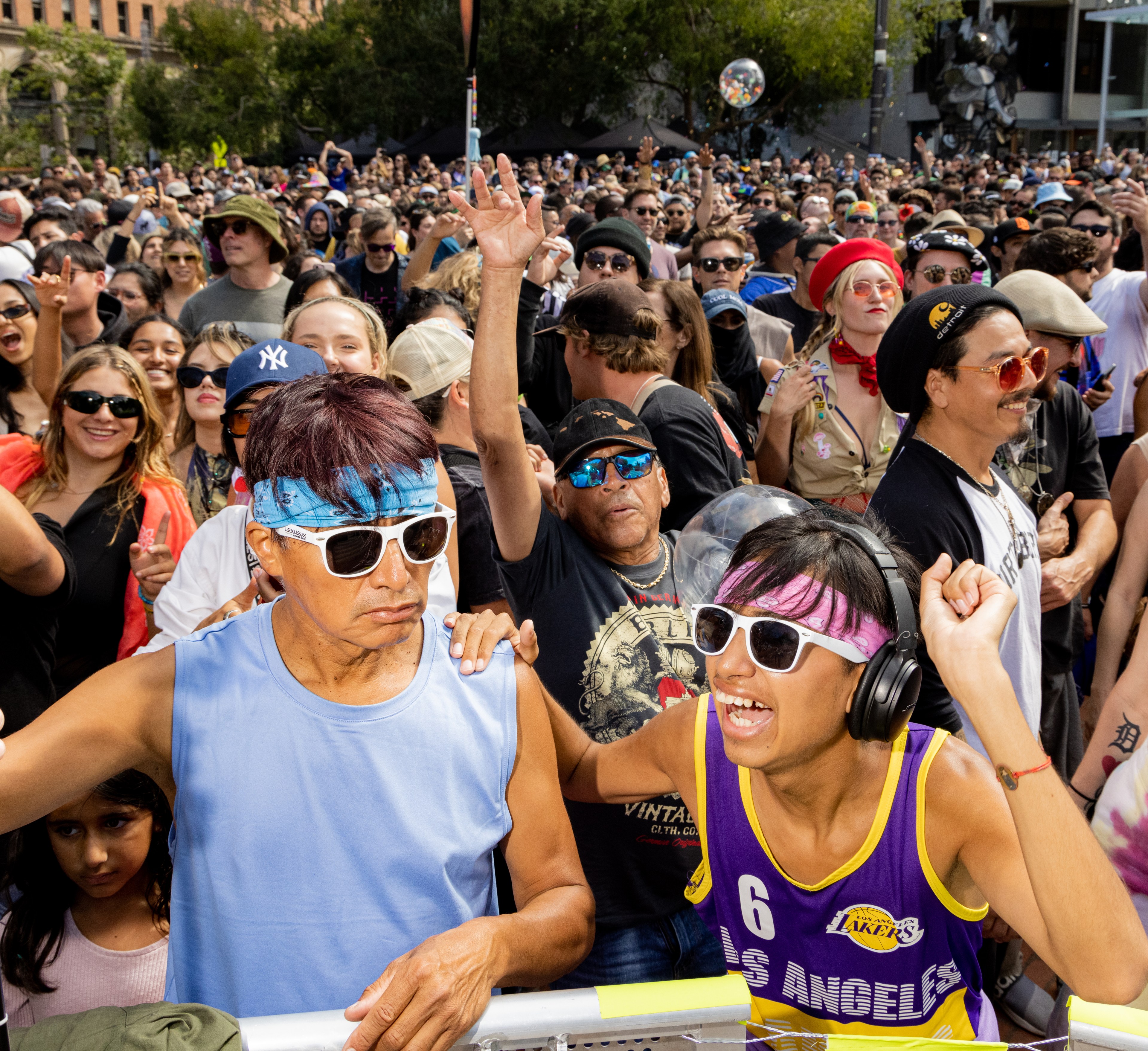 A diverse crowd enjoys an energetic outdoor event. People are smiling, wearing sunglasses, and dancing, with one wearing headphones and another in a Lakers jersey.