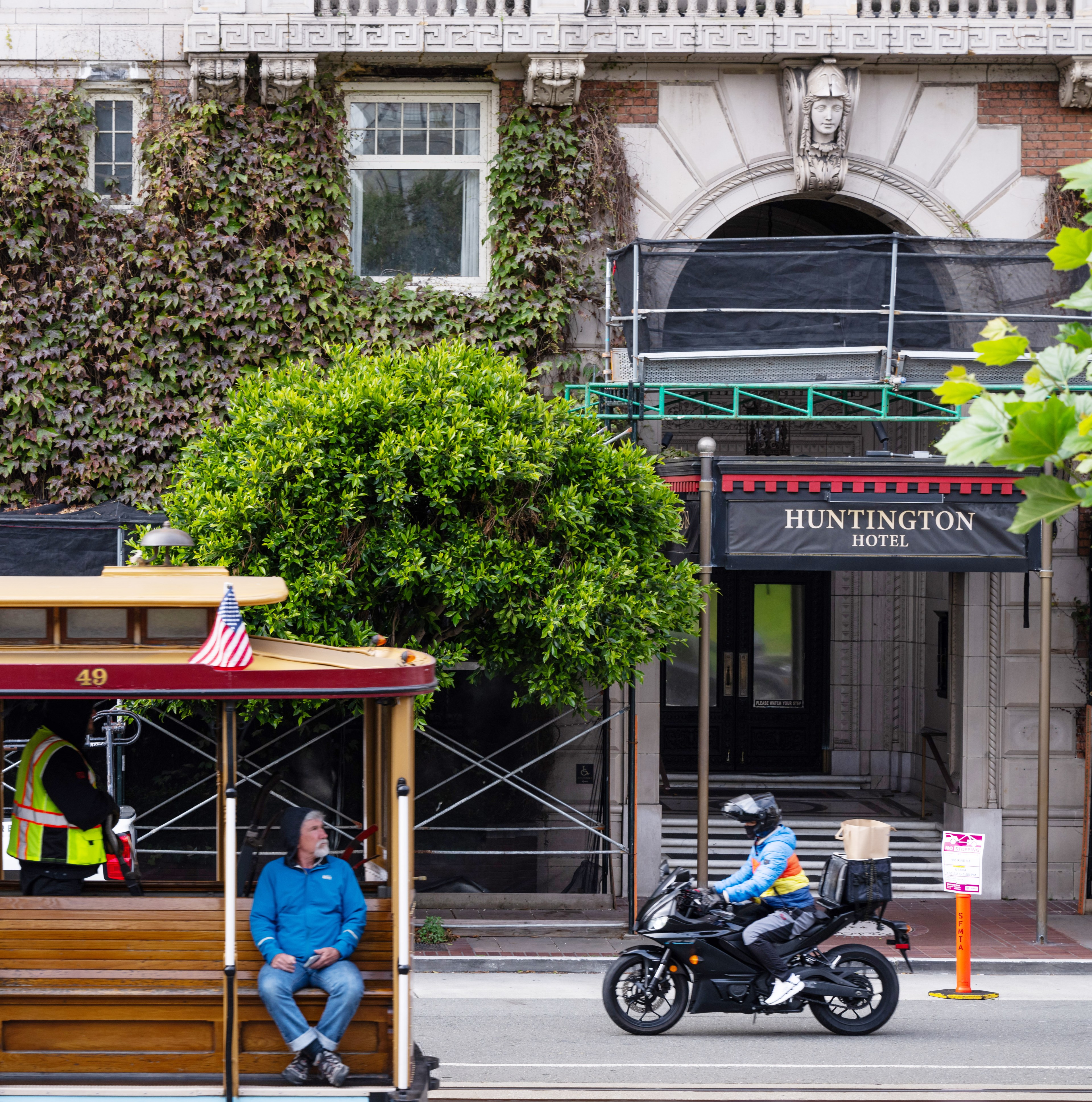 A man on a trolley car looks towards a motorcyclist riding in front of an entrance to the Huntington Hotel. The building is covered in ivy with scaffolding above the entrance.