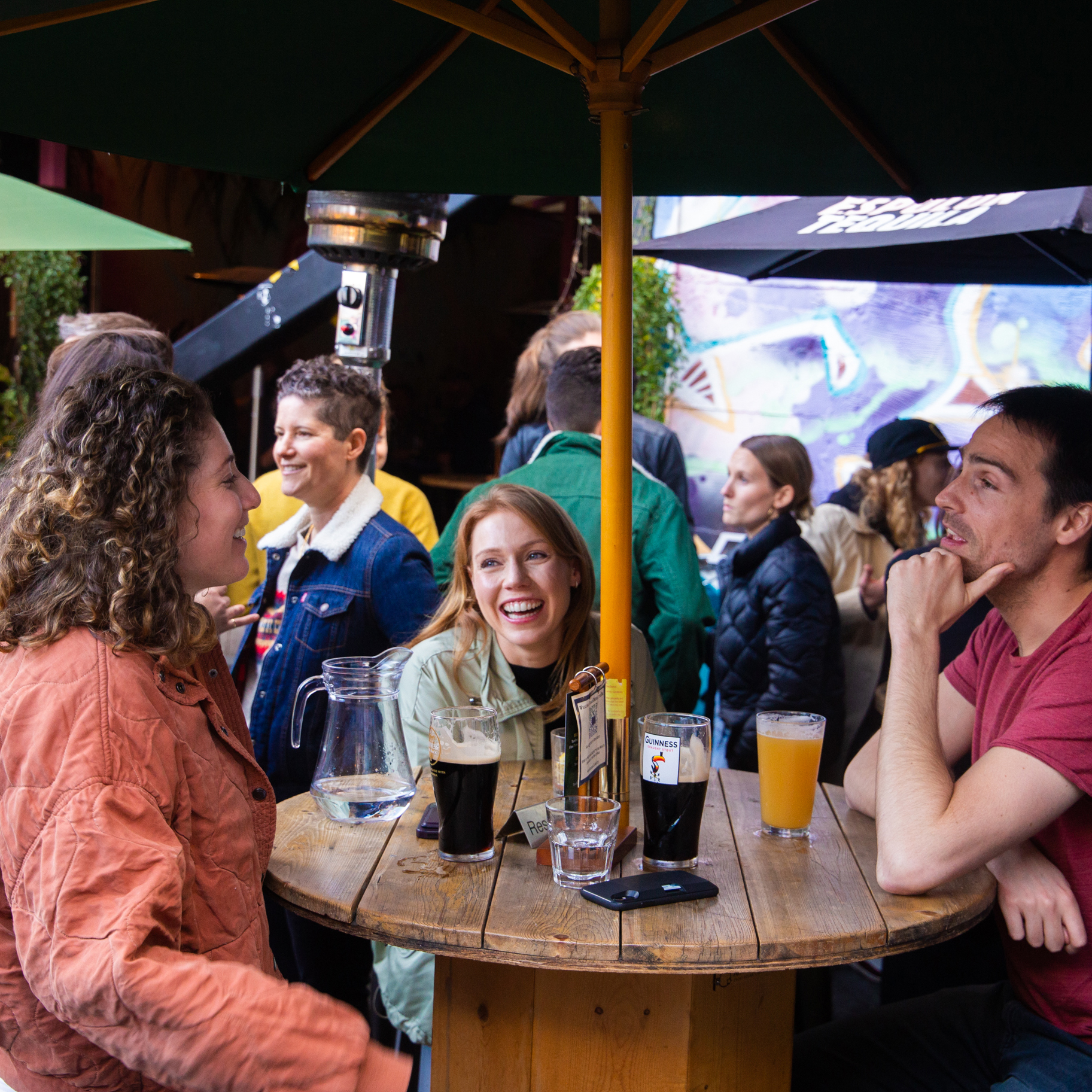 A group of people is gathered around a wooden table in an outdoor setting, drinking beer and talking, with a busy background filled with more people and vibrant graffiti.