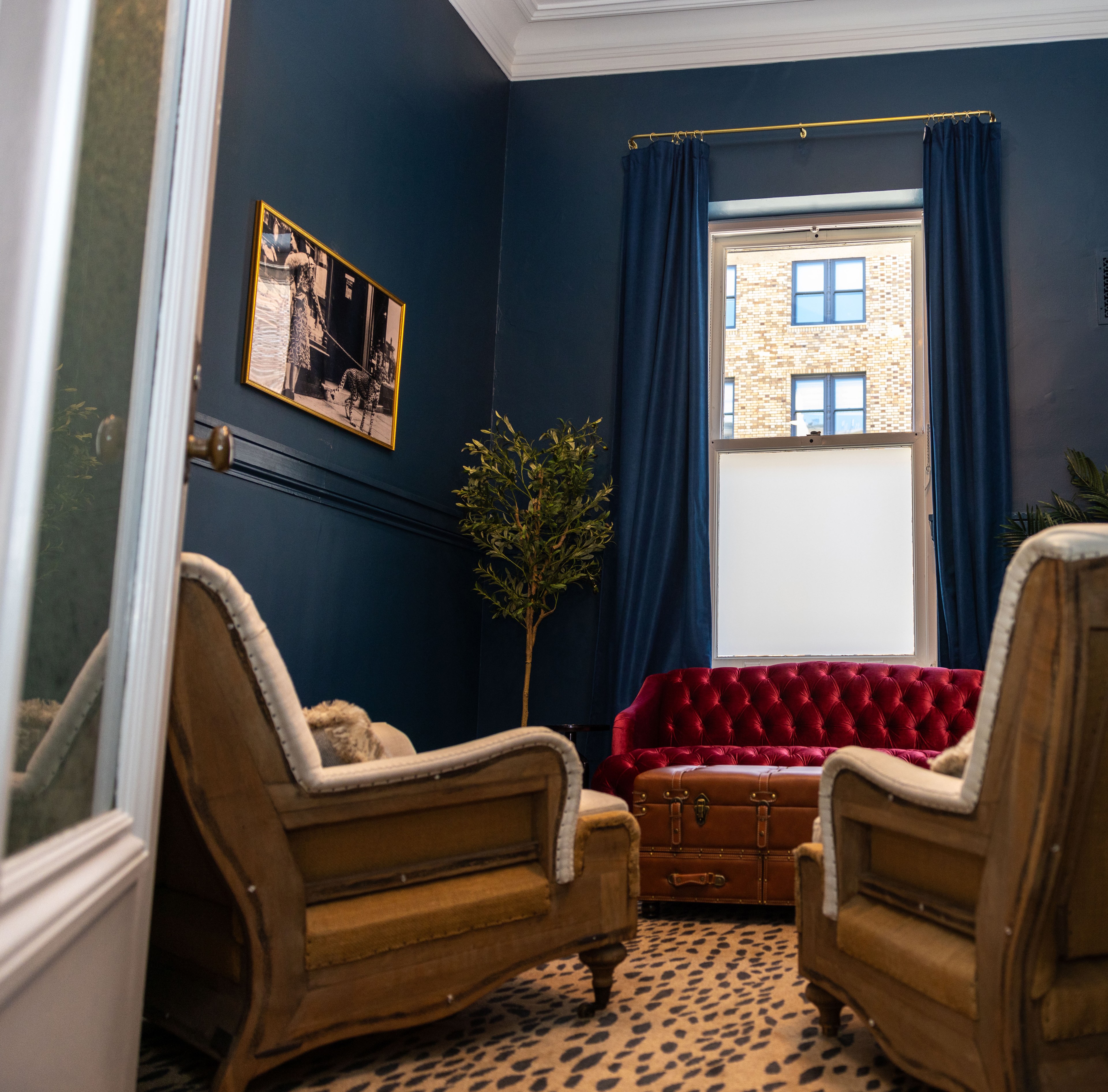 The room has deep blue walls, a red tufted sofa, two beige armchairs, and a window with blue curtains. A potted plant and a framed picture are on the wall.