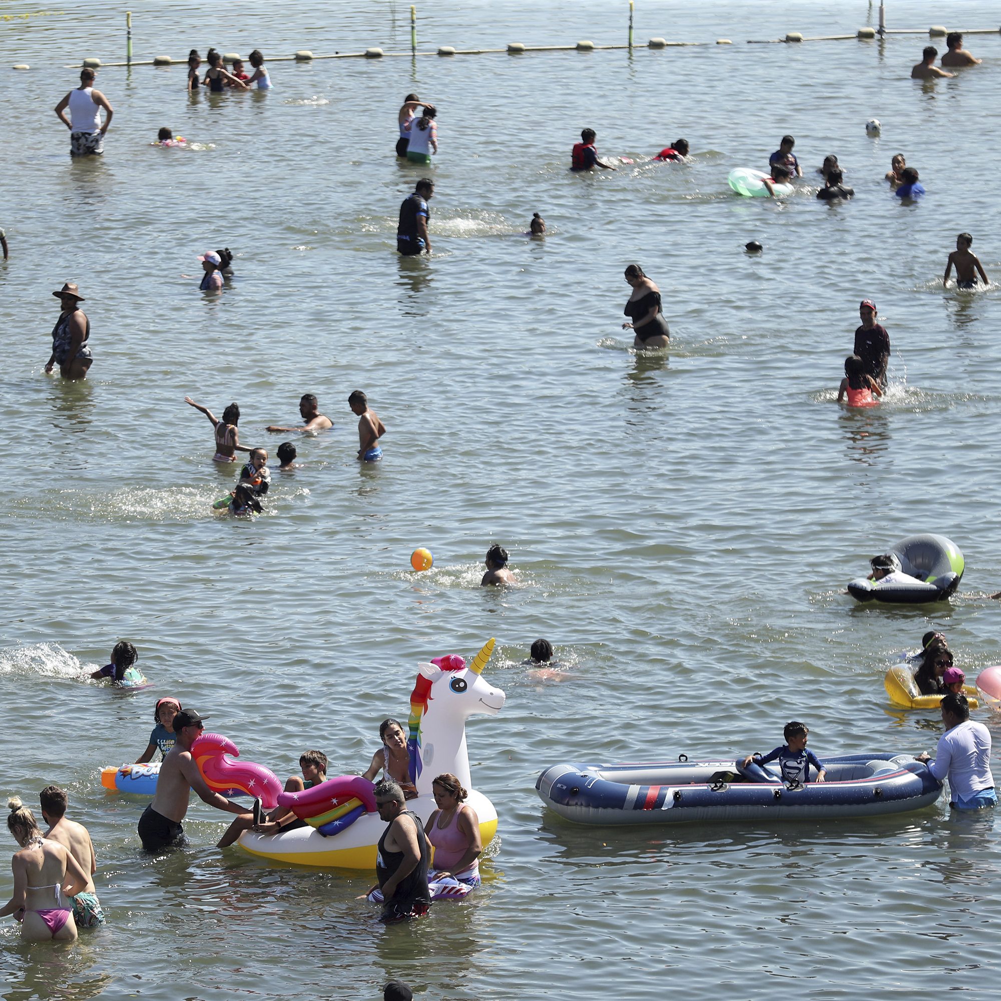People of all ages enjoy a sunny day wading and swimming in a lake. Some float on colorful inflatable rafts, including a unicorn, while others splash and play in the water.