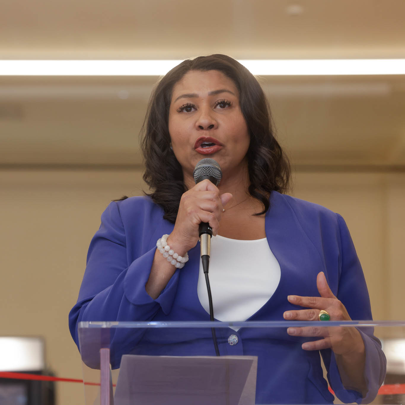 A woman in a blue blazer and white top is speaking into a microphone, standing behind a clear podium. She has dark wavy hair and is wearing a beaded bracelet.