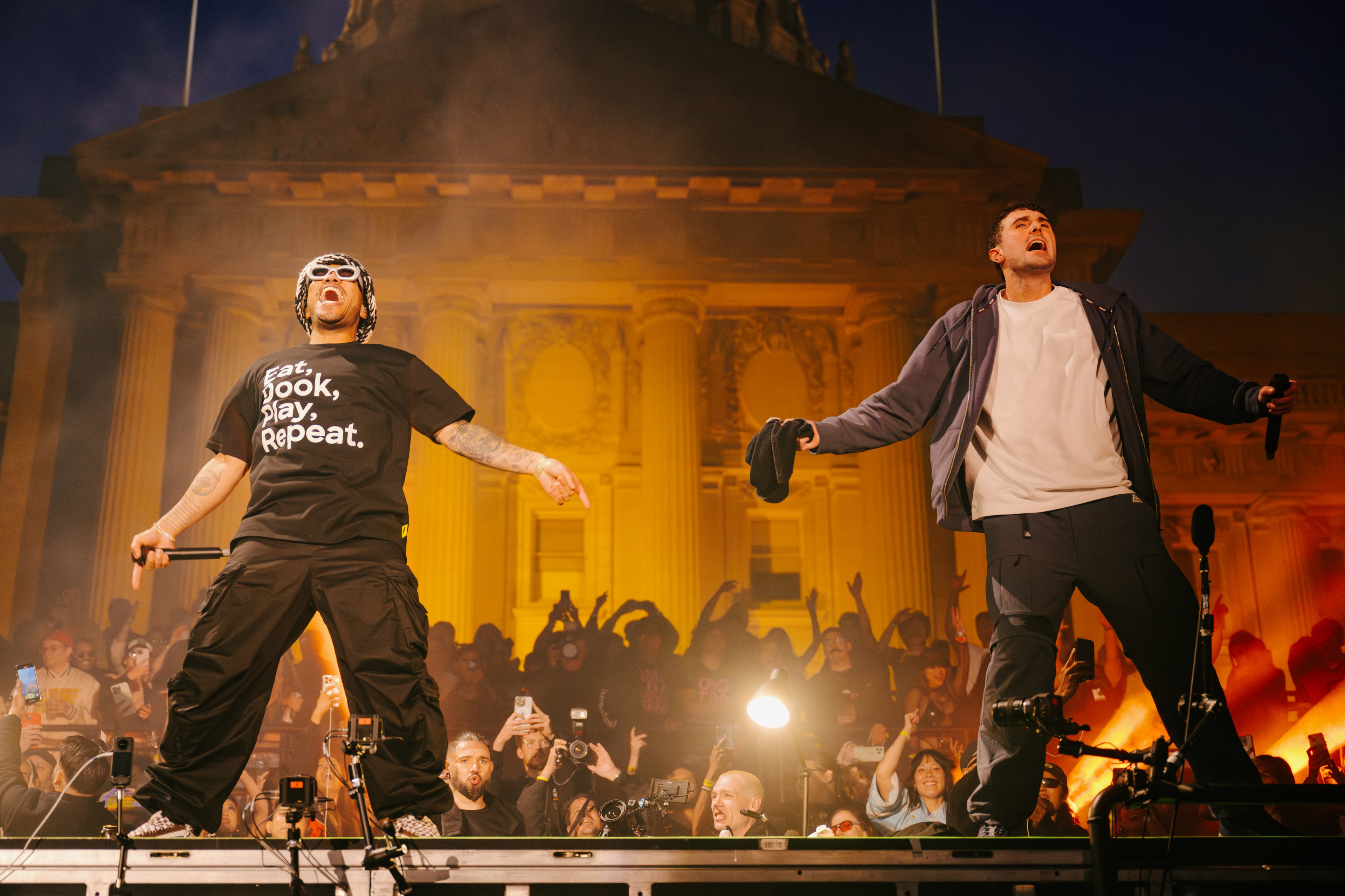 Two performers stand on stage, engaging with a lively crowd in front of an illuminated building. One wears sunglasses and a slogan tee, the other a jacket.