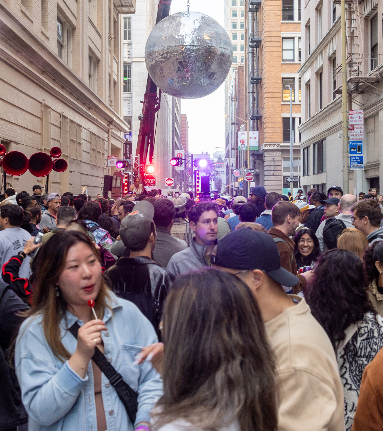 A large crowd gathers in a city street, observing a huge silver disco ball suspended from a crane. People are chatting, some are looking at the display, and there are lights and street signs visible.