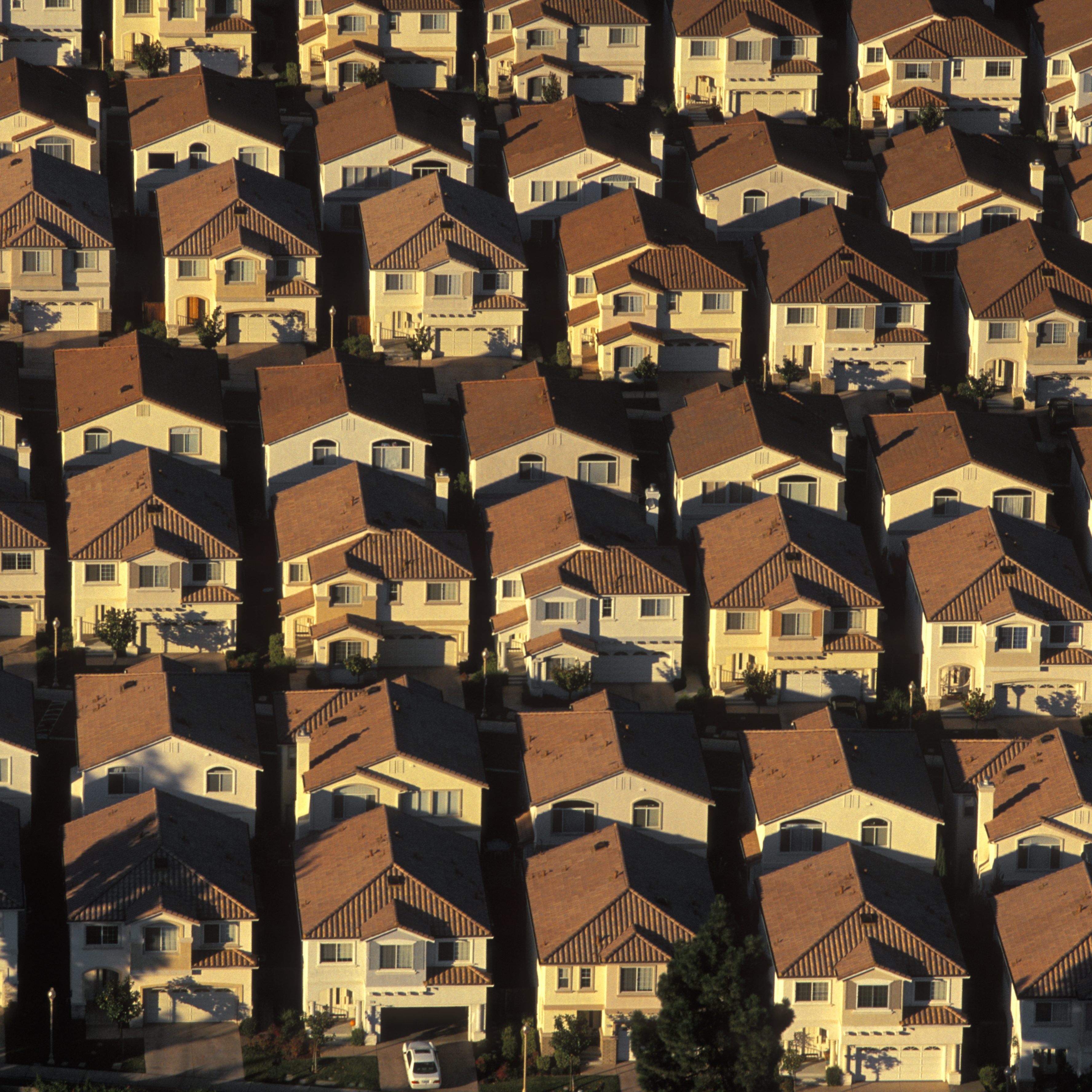 Rows of identical two-story houses with red-tiled roofs are closely packed together, creating a grid-like pattern, with shadows accentuating the uniformity.