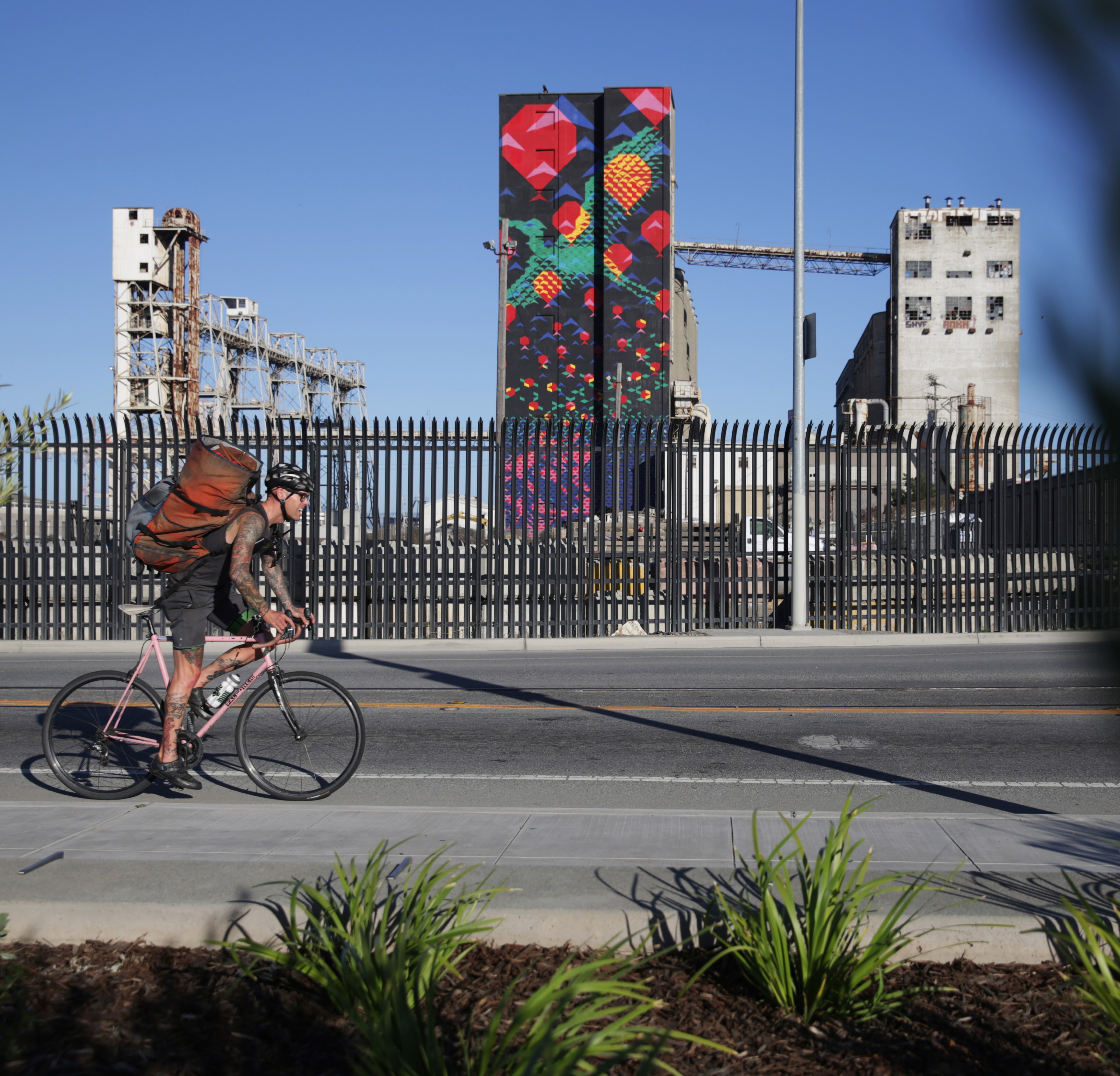 A cyclist with a backpack rides on a street next to a fence. Behind the fence are industrial buildings, including a tall, colorful mural painted with geometric shapes.