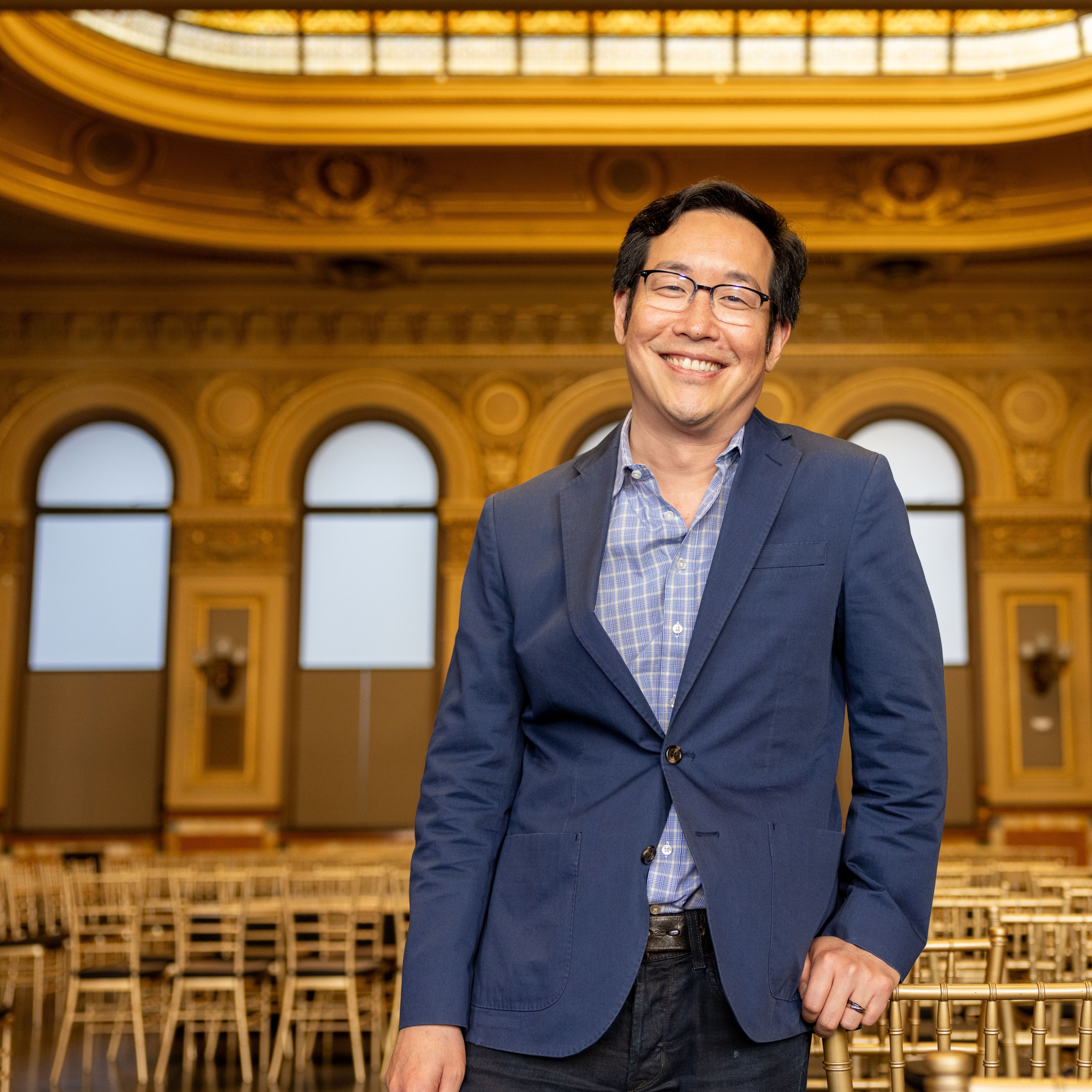 A smiling man in a blue blazer and glasses stands in an ornate room with arched windows and gold detailing, holding a chair in front of a row of empty chairs.