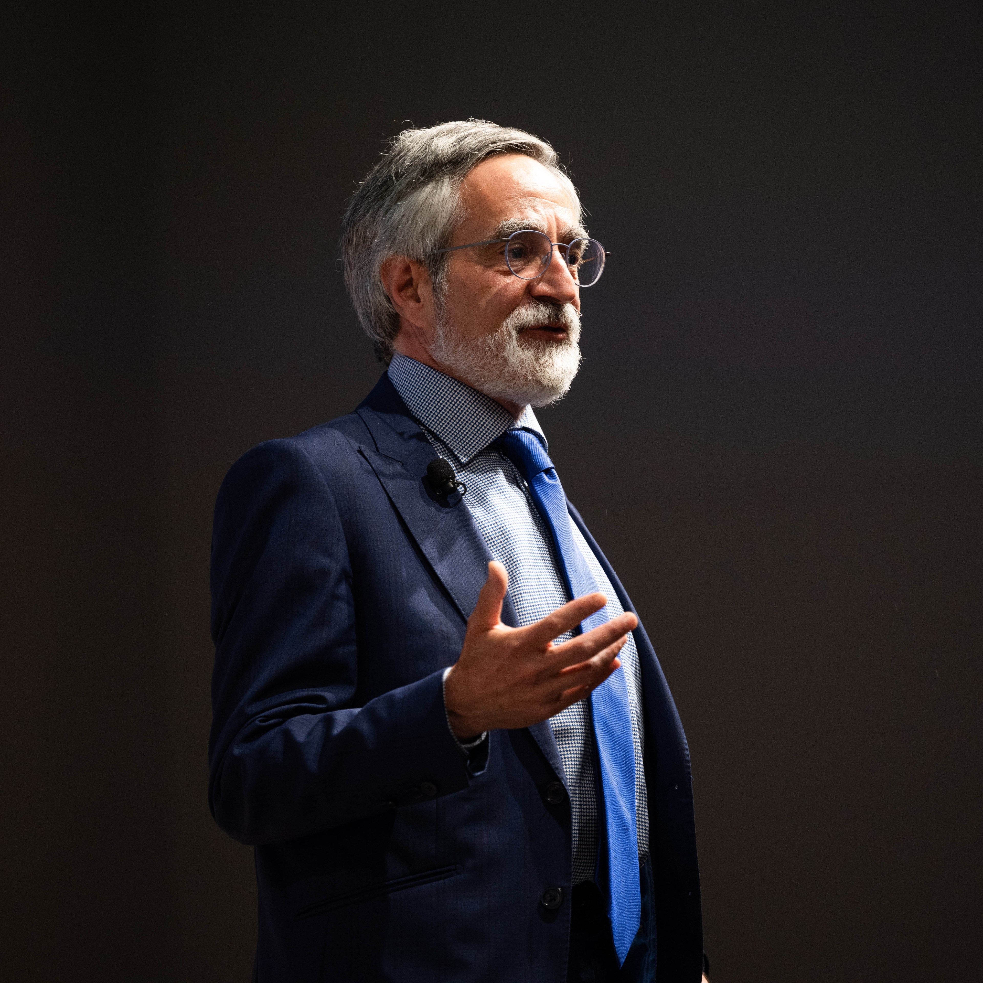 An older man with glasses and a beard is speaking, gesturing with his right hand. He is wearing a dark blue suit, a light blue shirt, and a blue tie, with a microphone attached.