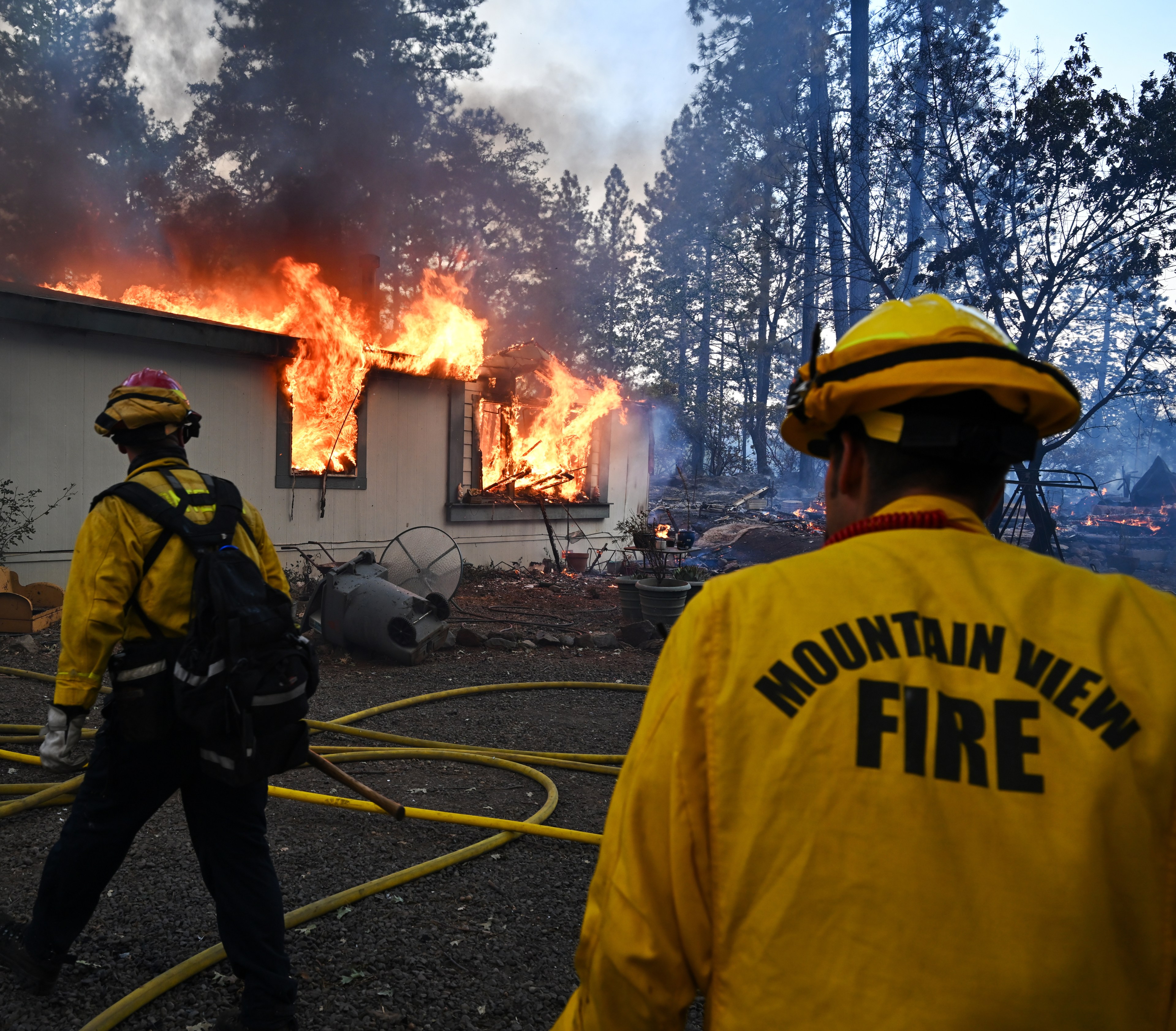 Two firefighters in yellow gear from Mountain View Fire Department stand near a house engulfed in bright orange flames, with hoses on the ground amidst dense trees.