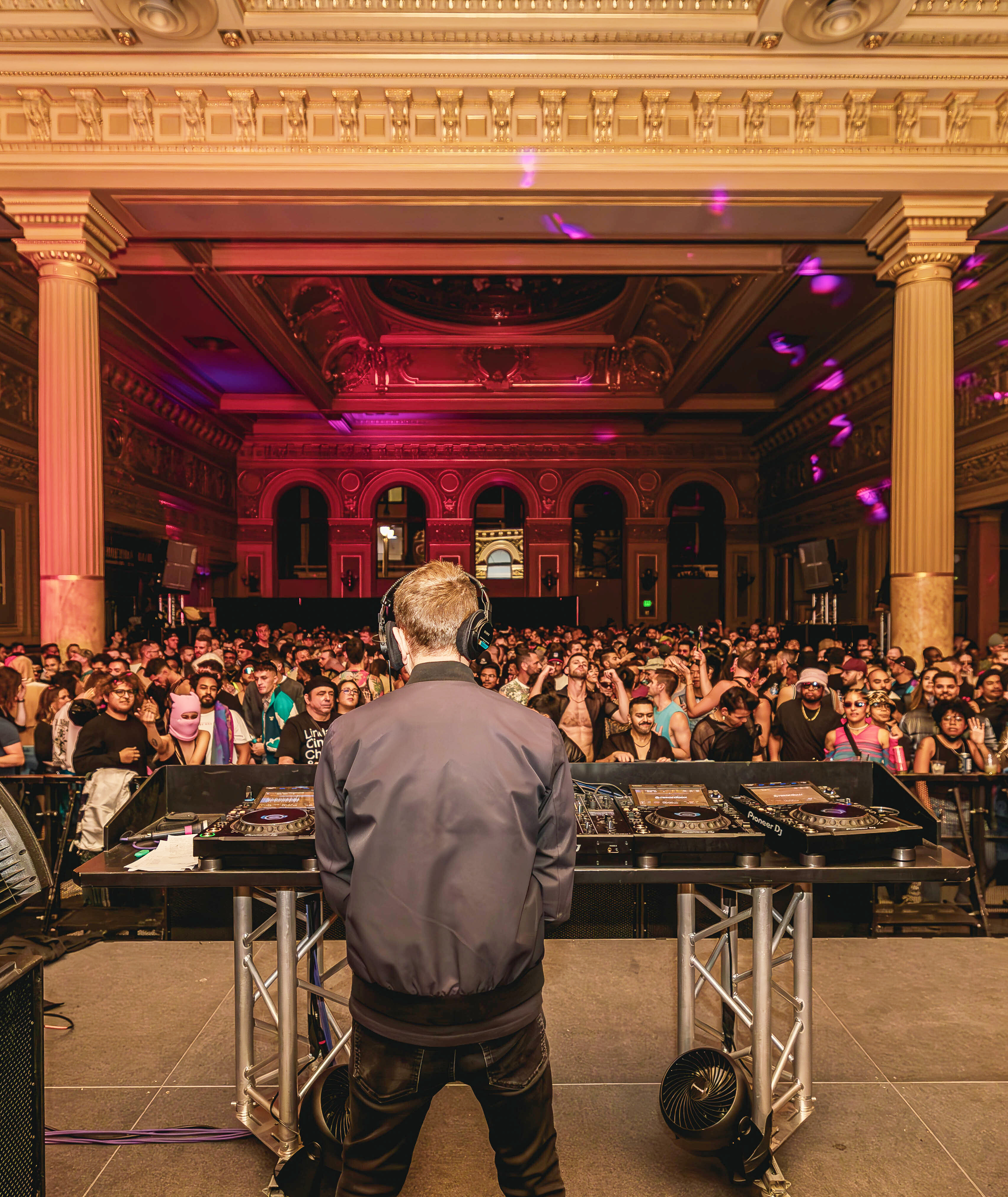 A DJ performs in a grand, ornate hall with high ceilings, columns, and elaborate decor. The crowd is energetic and diverse, filling the room with excitement.