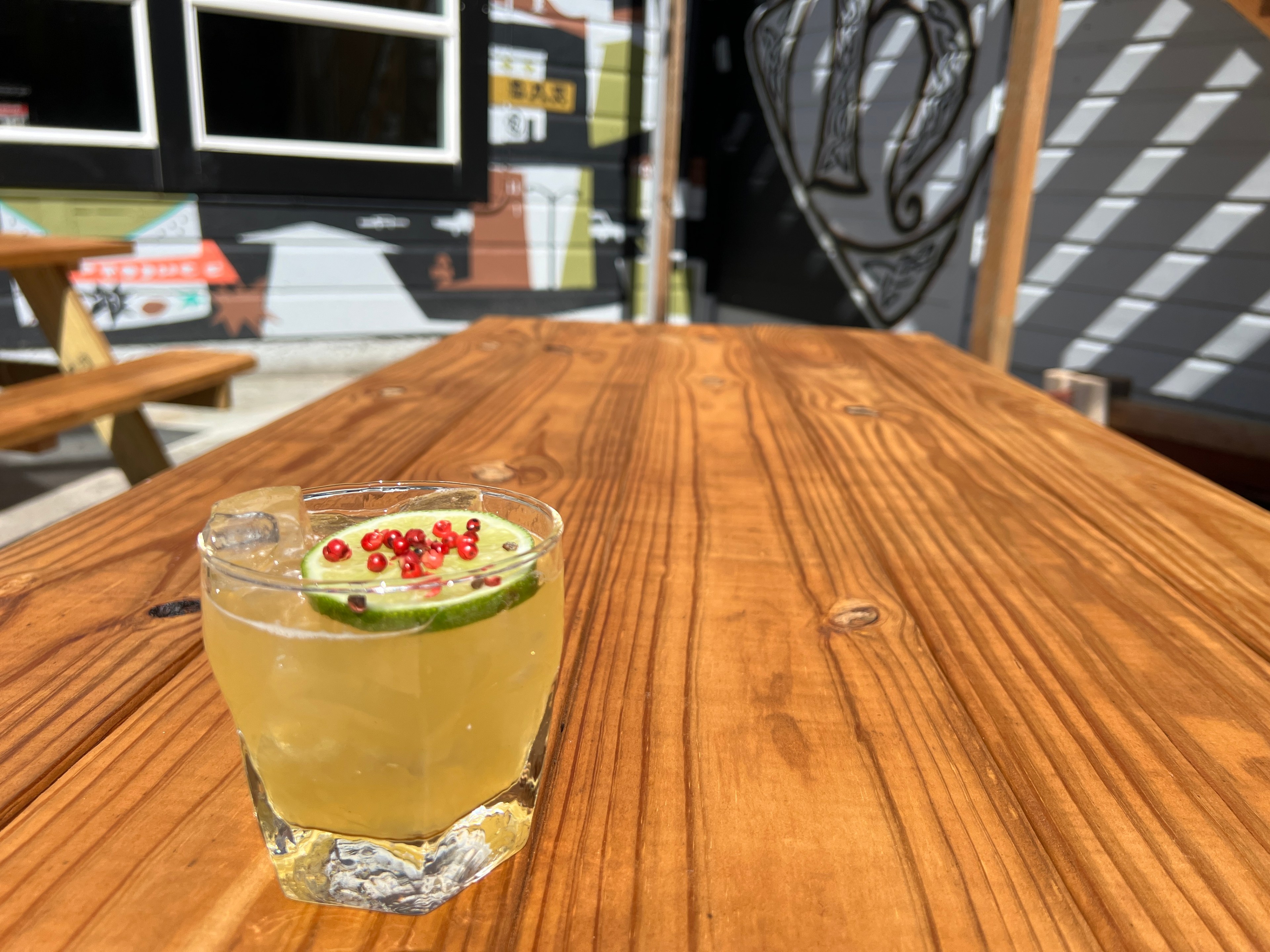 A chilled drink garnished with a lime slice and red peppercorns sits on a wooden picnic table outdoors, with colorful building murals in the background.