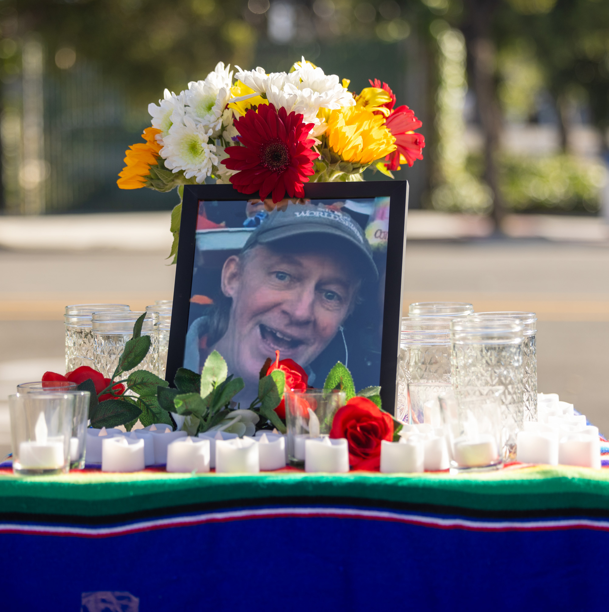 A framed photo of a smiling man is set on a colorful cloth surrounded by candles, flowers, and glass jars, in an outdoor setting.