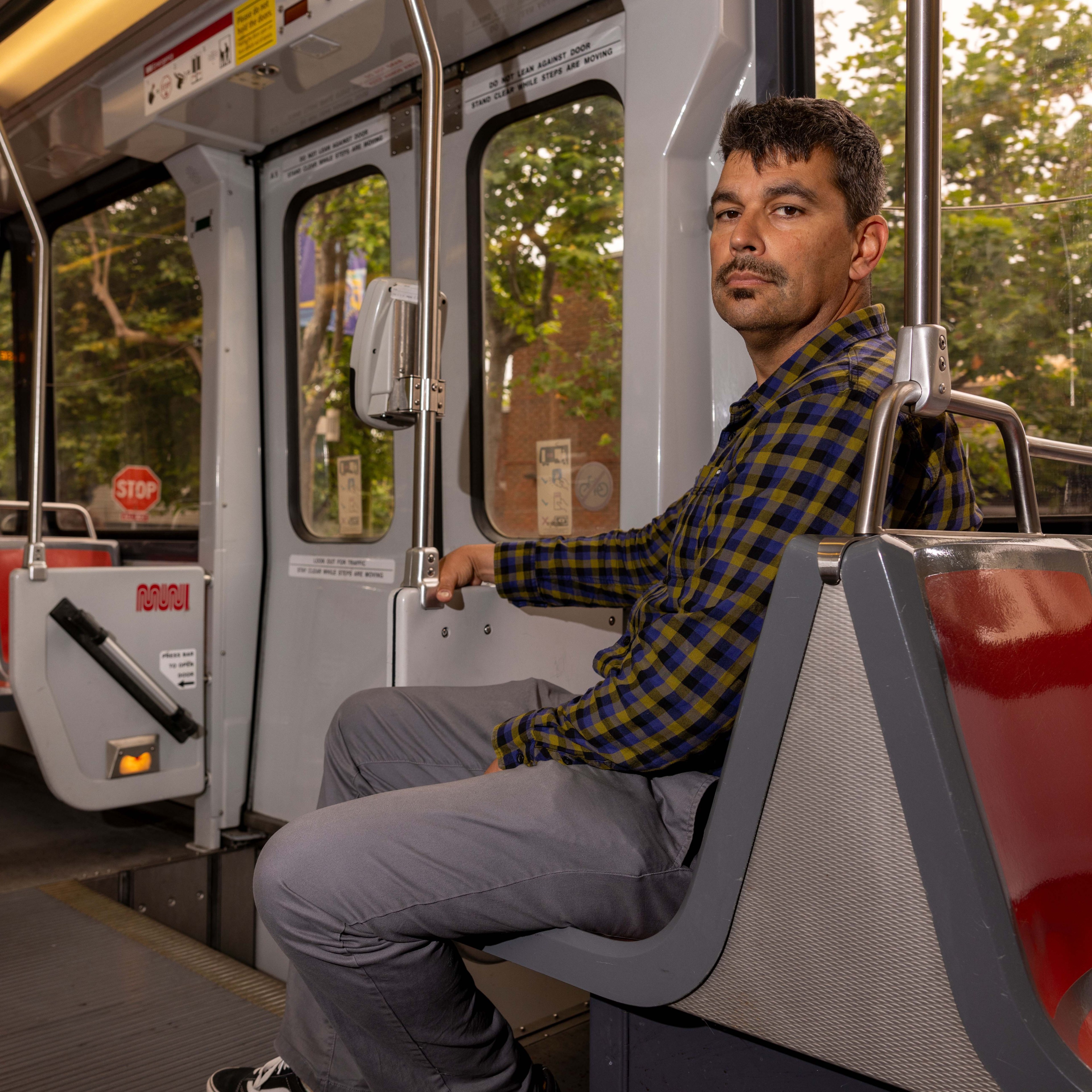 A man with short hair and a mustache, wearing a plaid shirt and gray pants, sits on a public transit seat.