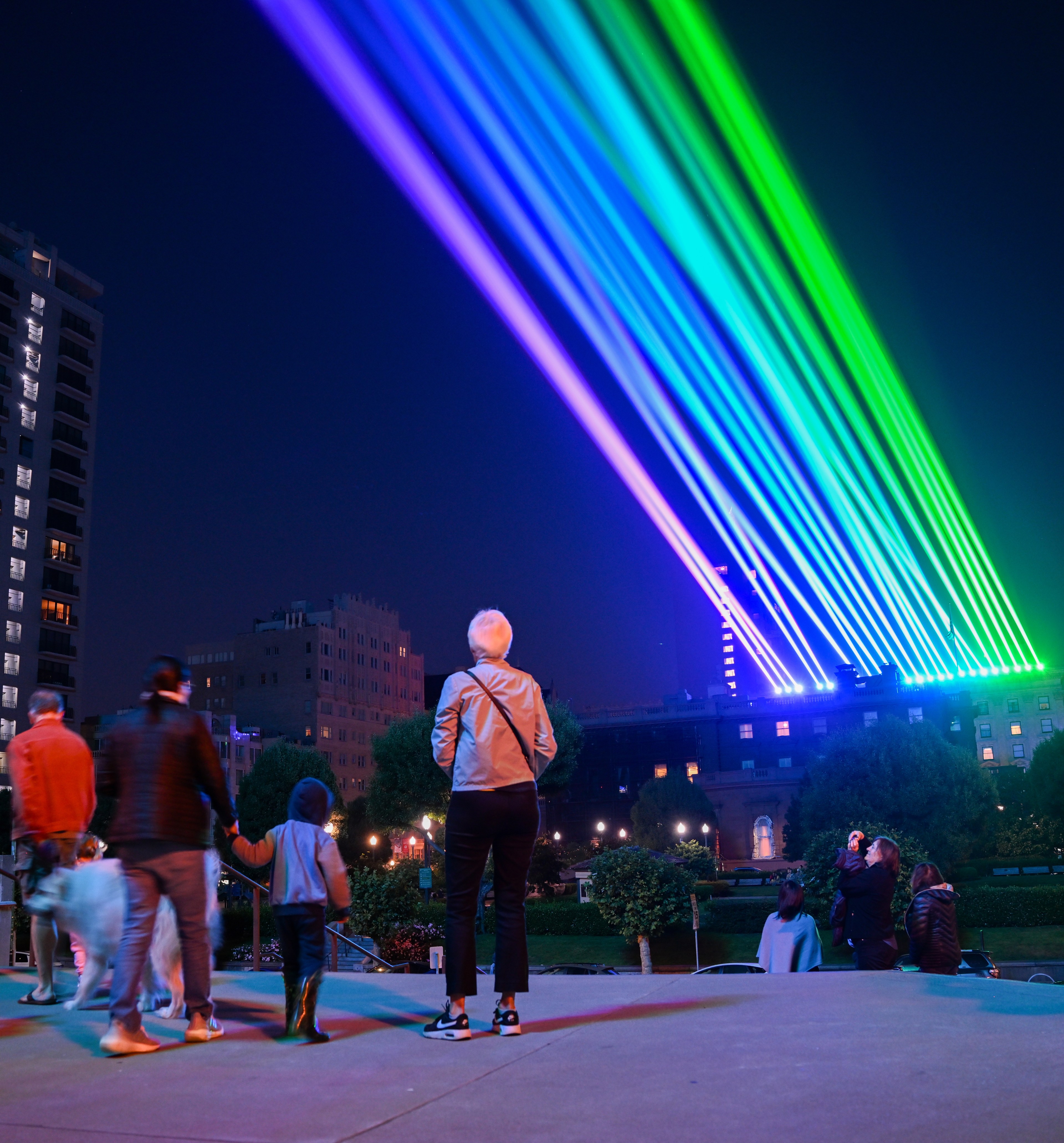 Several people stand in a nighttime urban park, admiring vibrant rainbow-colored laser beams projected into the sky from nearby buildings.