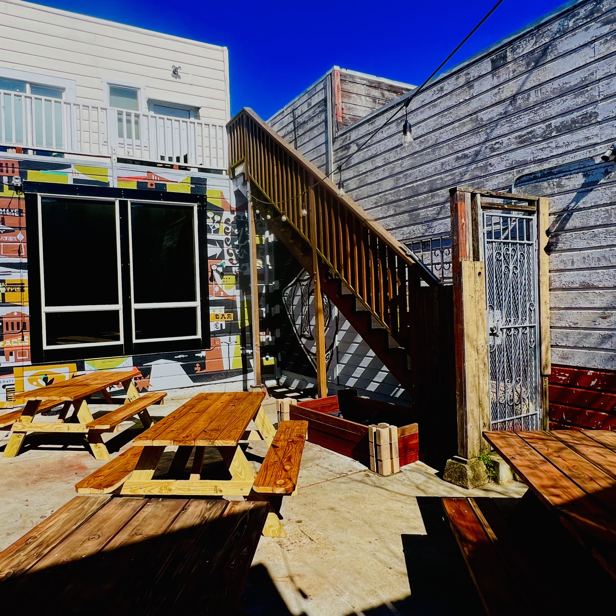 The image shows a courtyard with wooden picnic tables, a colorful mural, and a wooden staircase leading to an upper level. A blue sky is visible above.