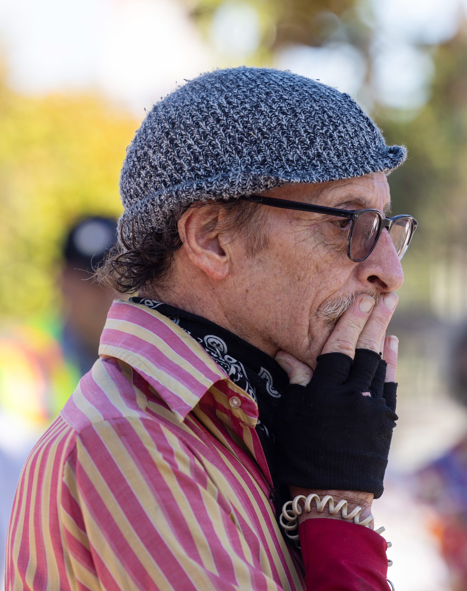 A man with glasses, a gray knit cap, and a striped shirt touches his face, wearing fingerless gloves and a black neck scarf, with a blurred outdoor background.