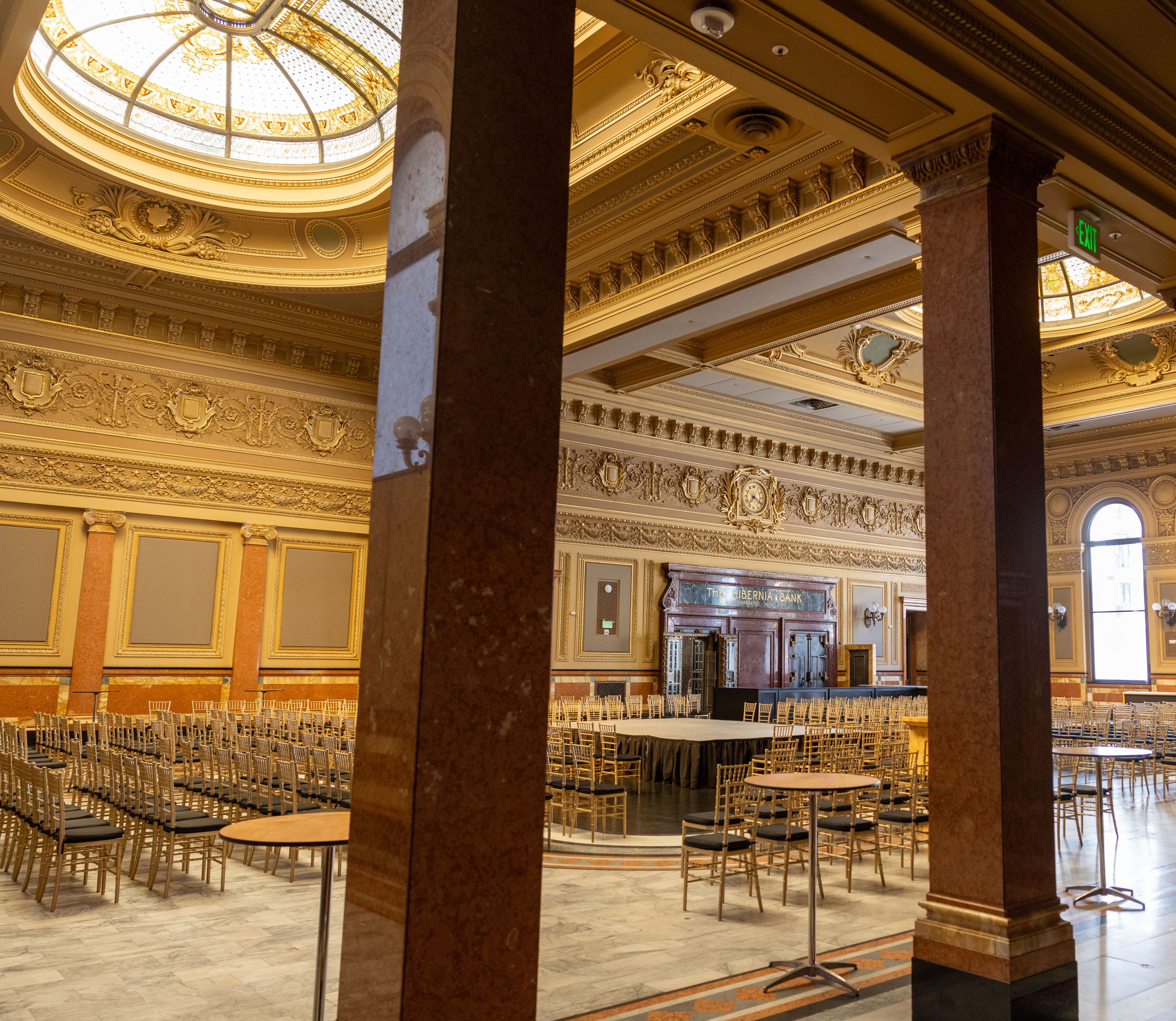 The image shows an ornate hall with a domed skylight, detailed golden moldings, rows of gold chairs, and a raised stage area. The walls are richly decorated with intricate designs.