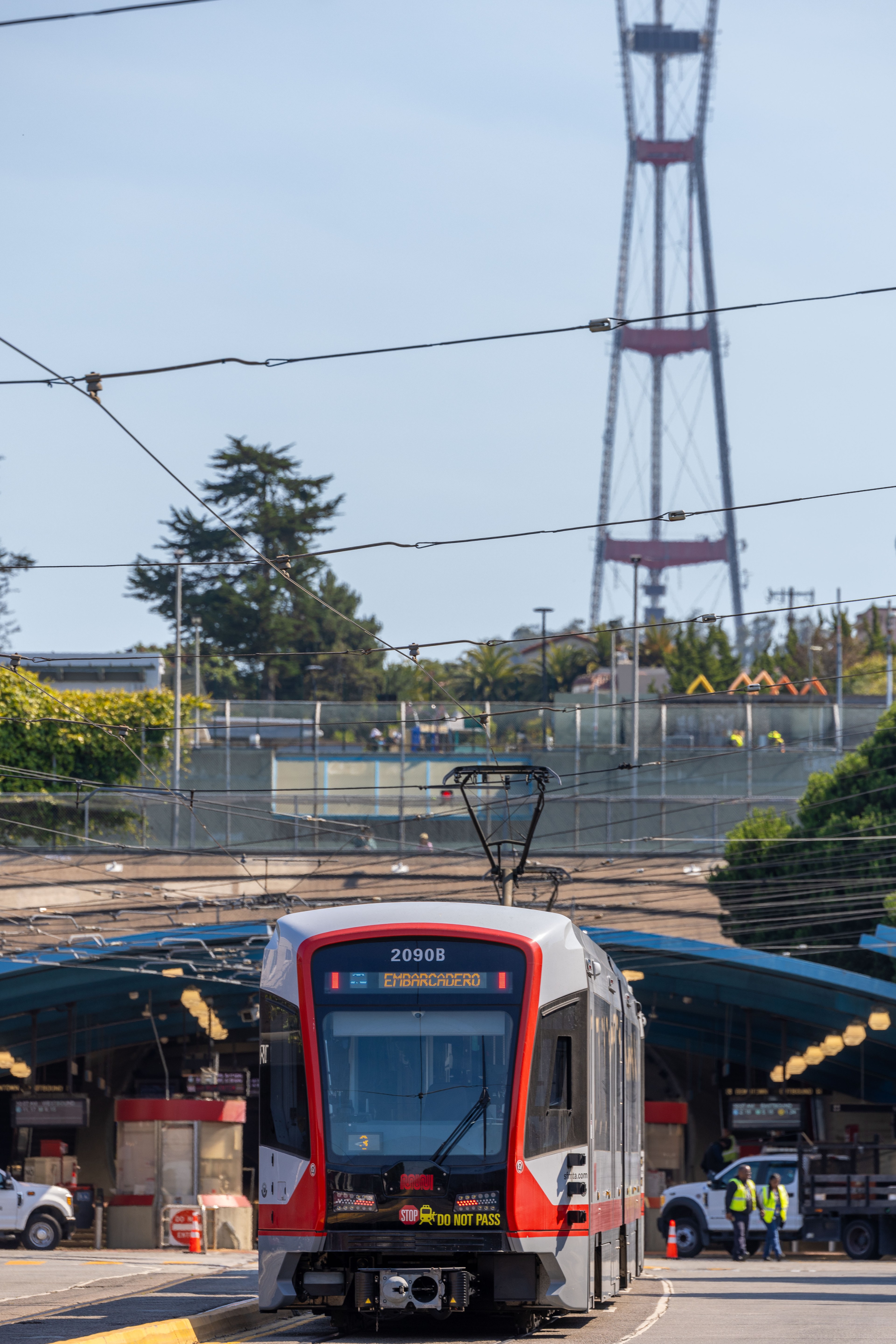 A modern red and white tram labeled &quot;2090B&quot; is on tracks with overhead wires, with a distant tall tower and trees visible in the background.